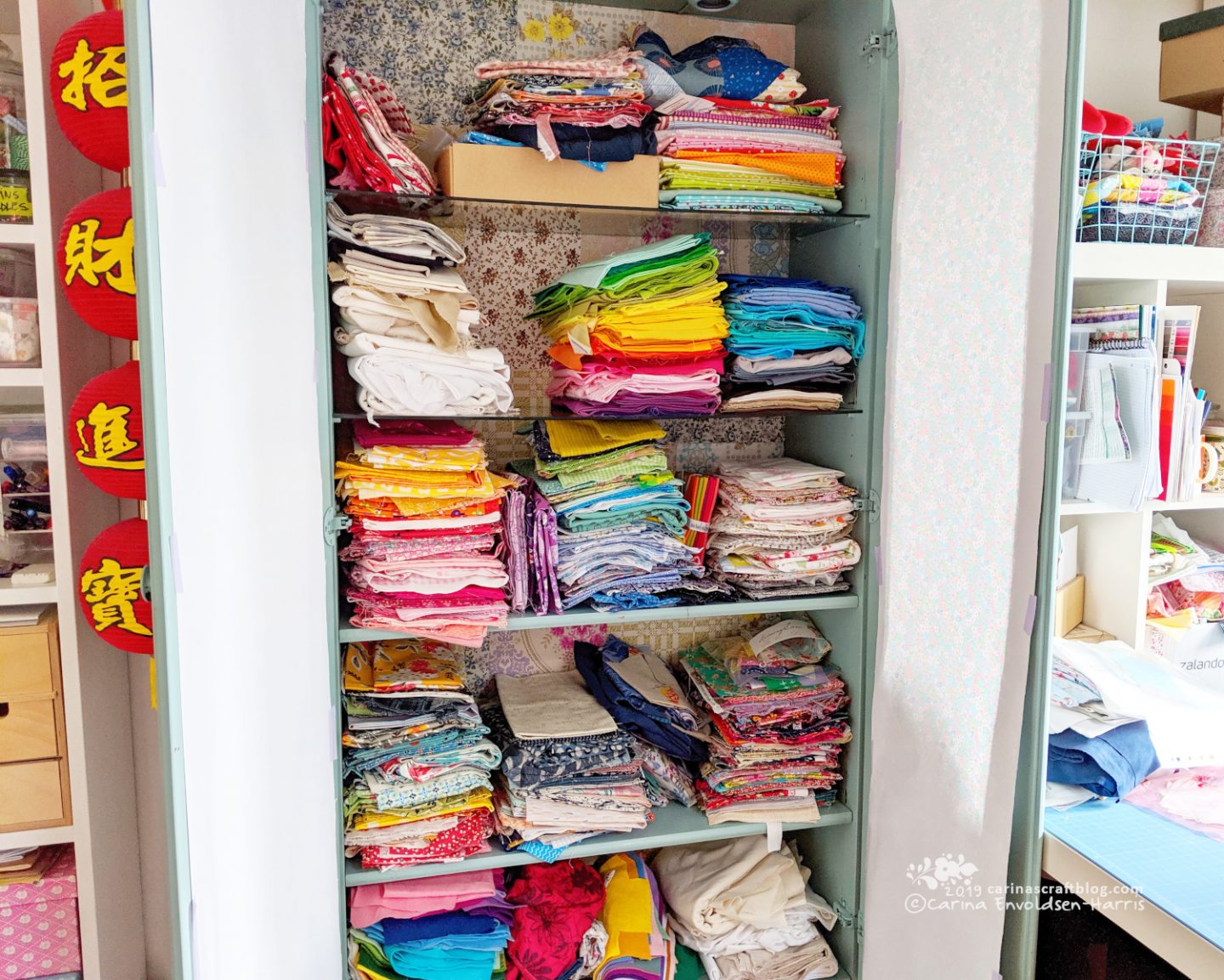 A tall cabinet with glass shelves, doors open so tidy stacks of fabric can be seen.