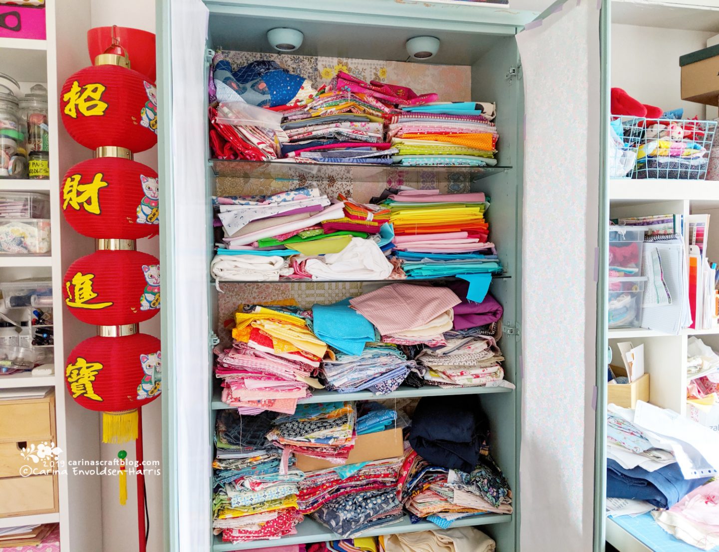 A tall cabinet with glass shelves, doors open so messy stacks of fabric can be seen.