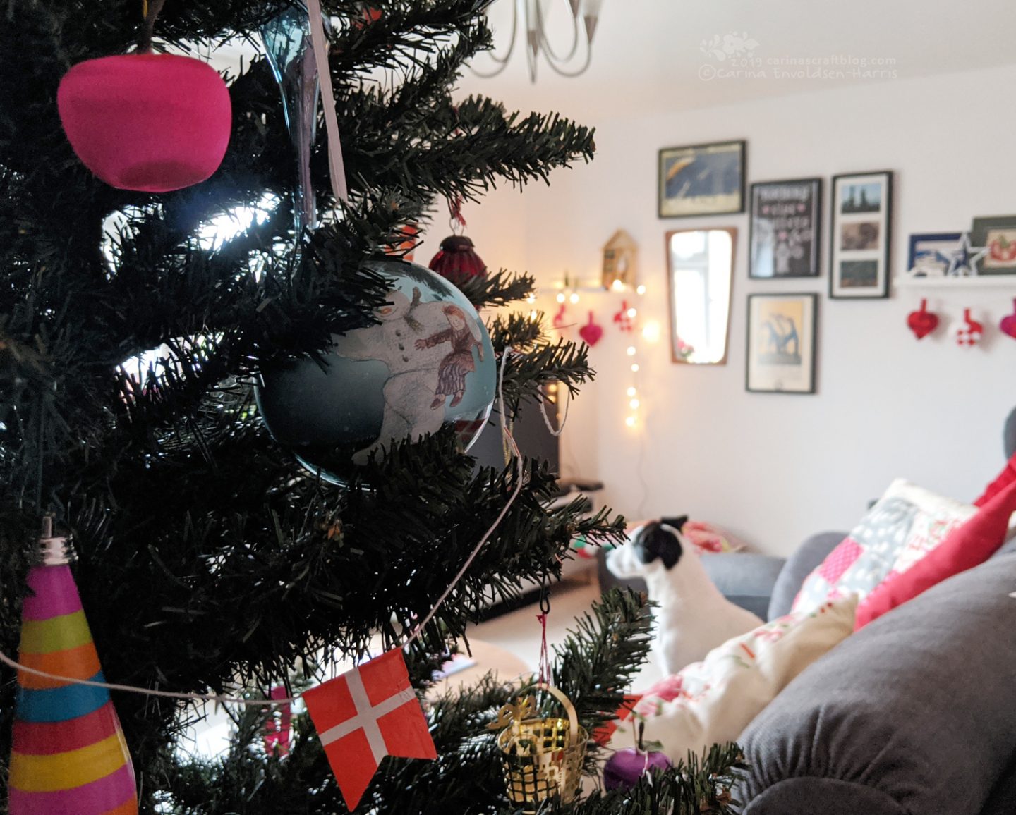 Decorated Christmas tree in the foreground, in teh background a picture wall, and a dog on a sofa.