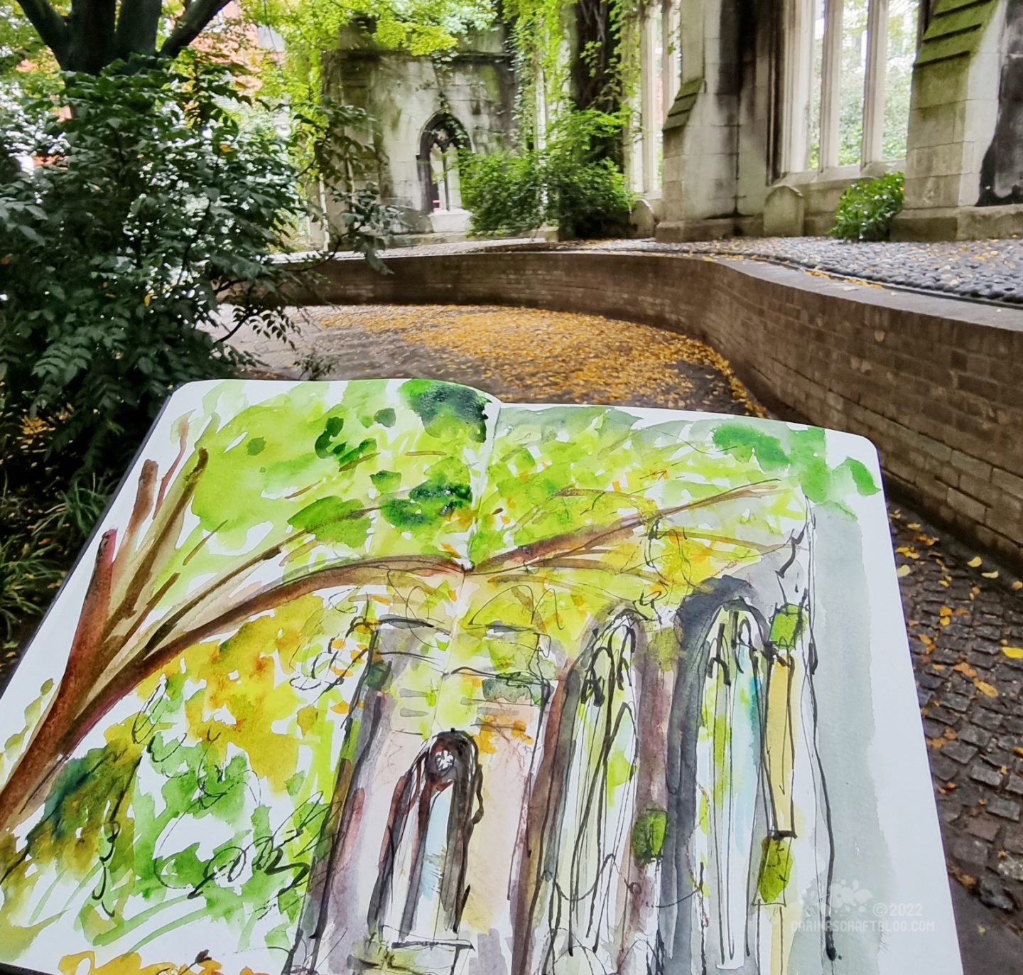 An open sketchbook held up in front of the subject on the pages: the ruins of a church overgrown with plants.