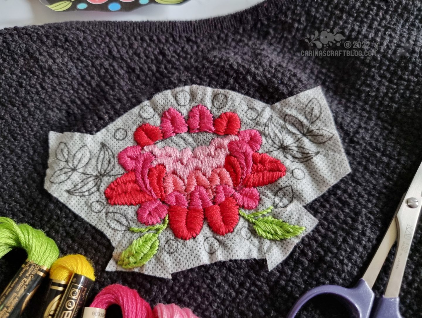 Partially embroidered folk art style flower in pink and red on a black cardigan.