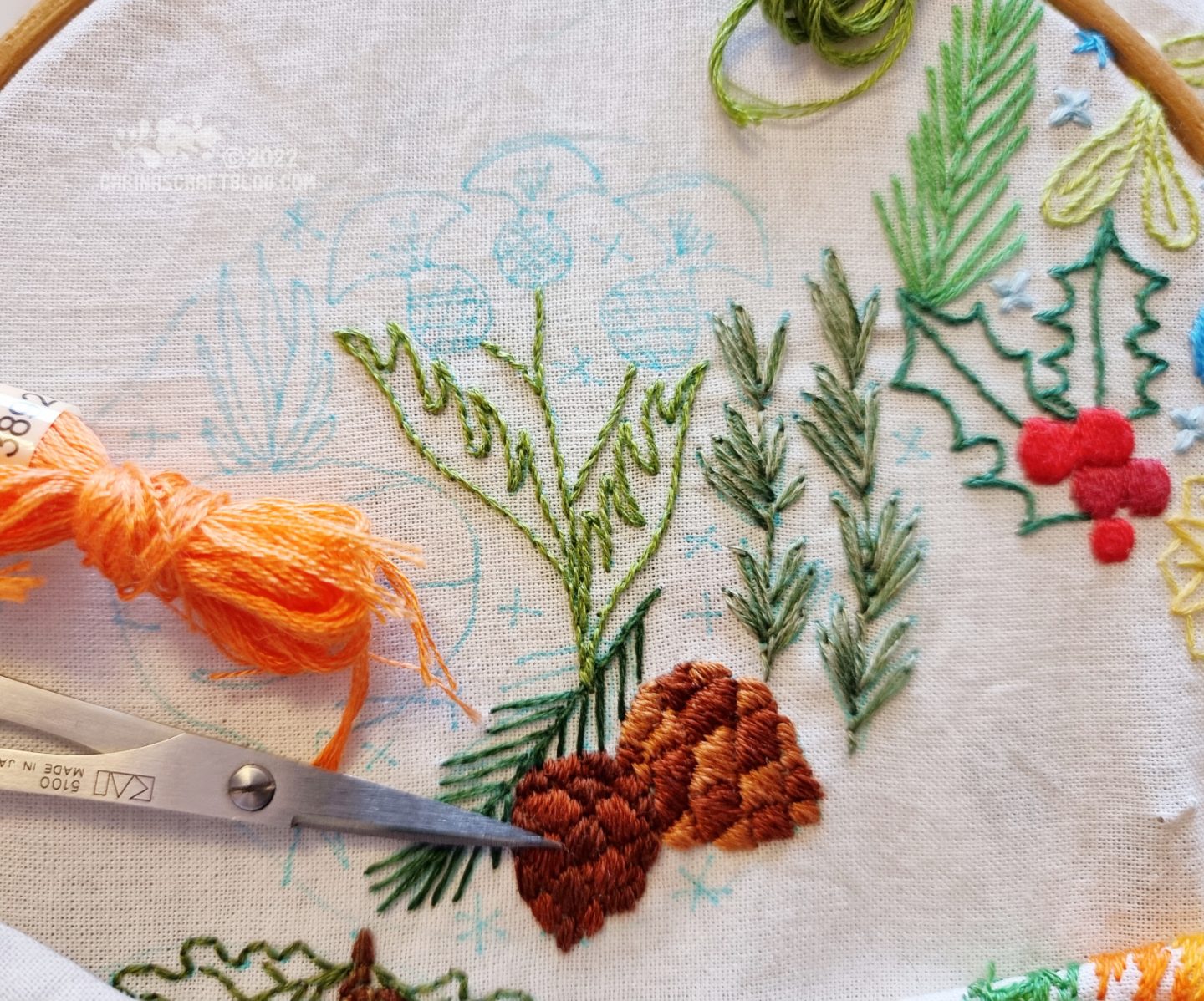 Closeup of embroidery on white fabric. Two pinecones are embroidered at the bottom, with rosemary next to them.