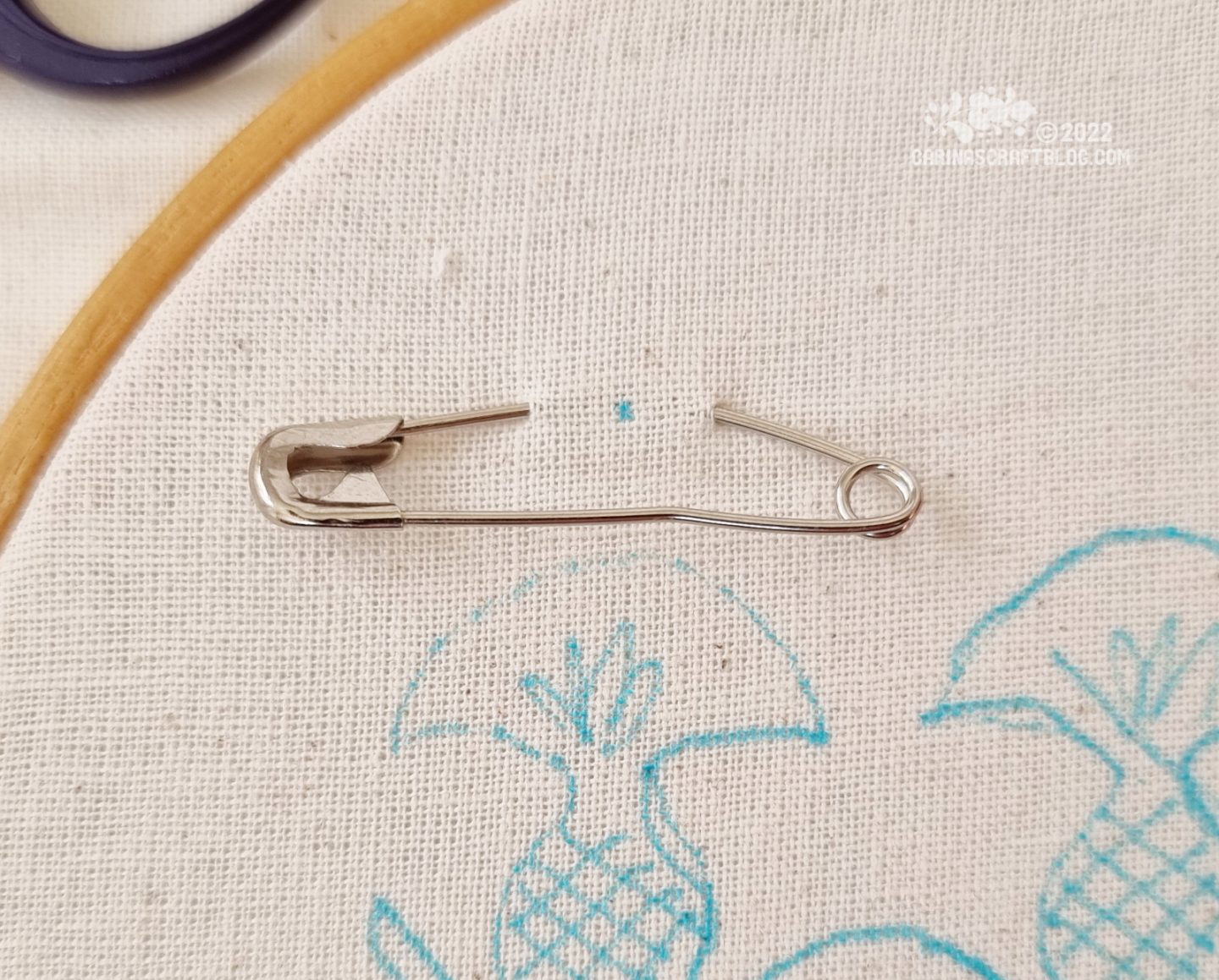 Closeup of a safety pin attached to white fabric in an embroidery hoop.