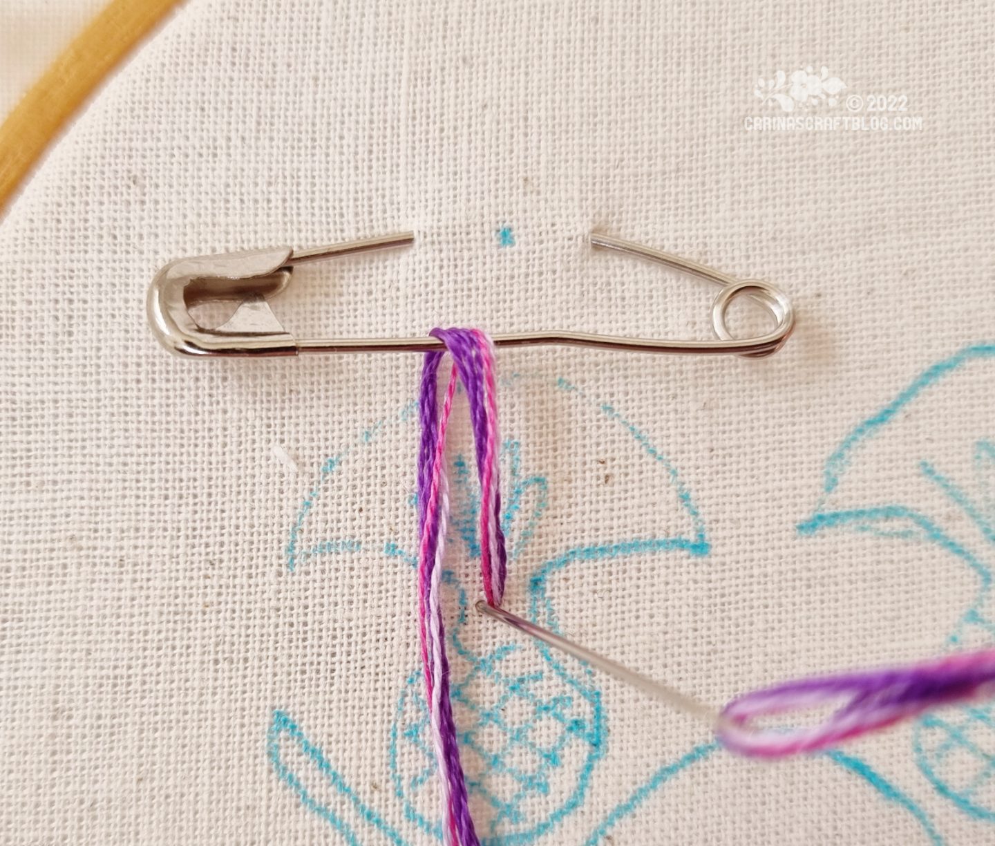 Closeup of a safety pin attached to white fabric. Embroidery thread in purple colours has been looped through the safety pin.