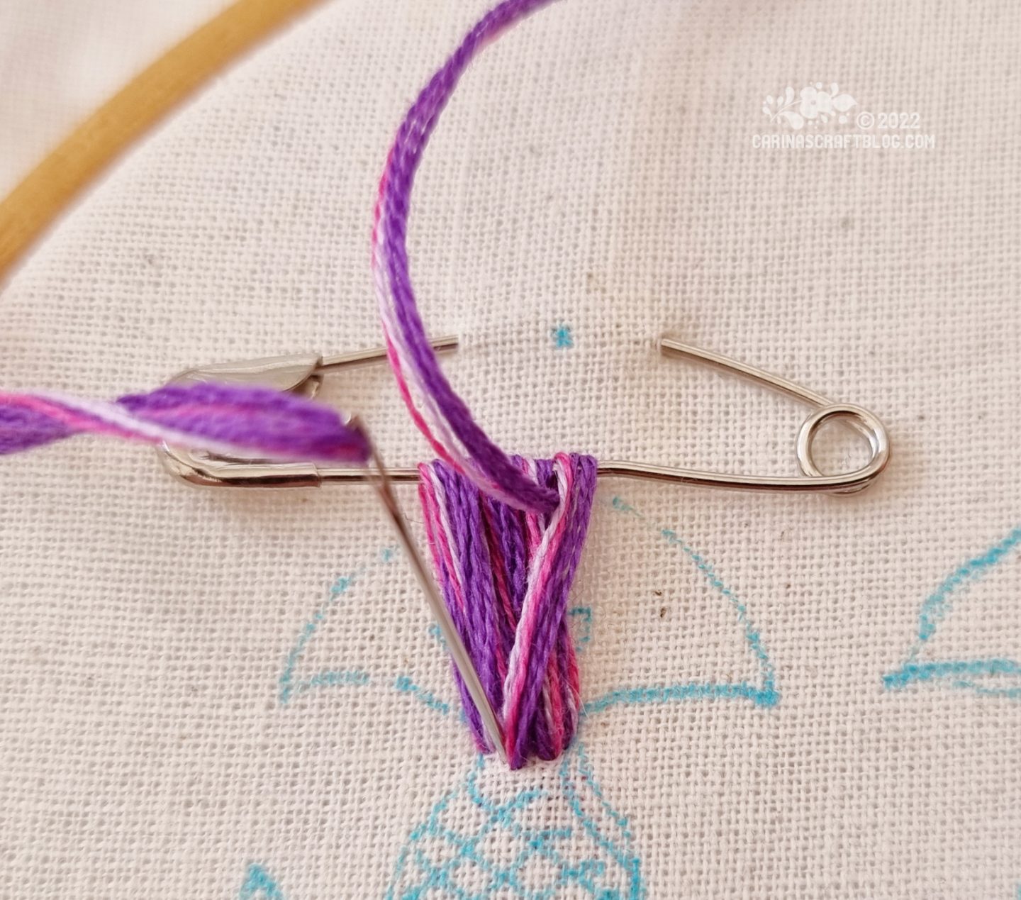Closeup of a safety pin attached to white fabric. Embroidery thread in purple colours has been looped through the safety pin several times.
