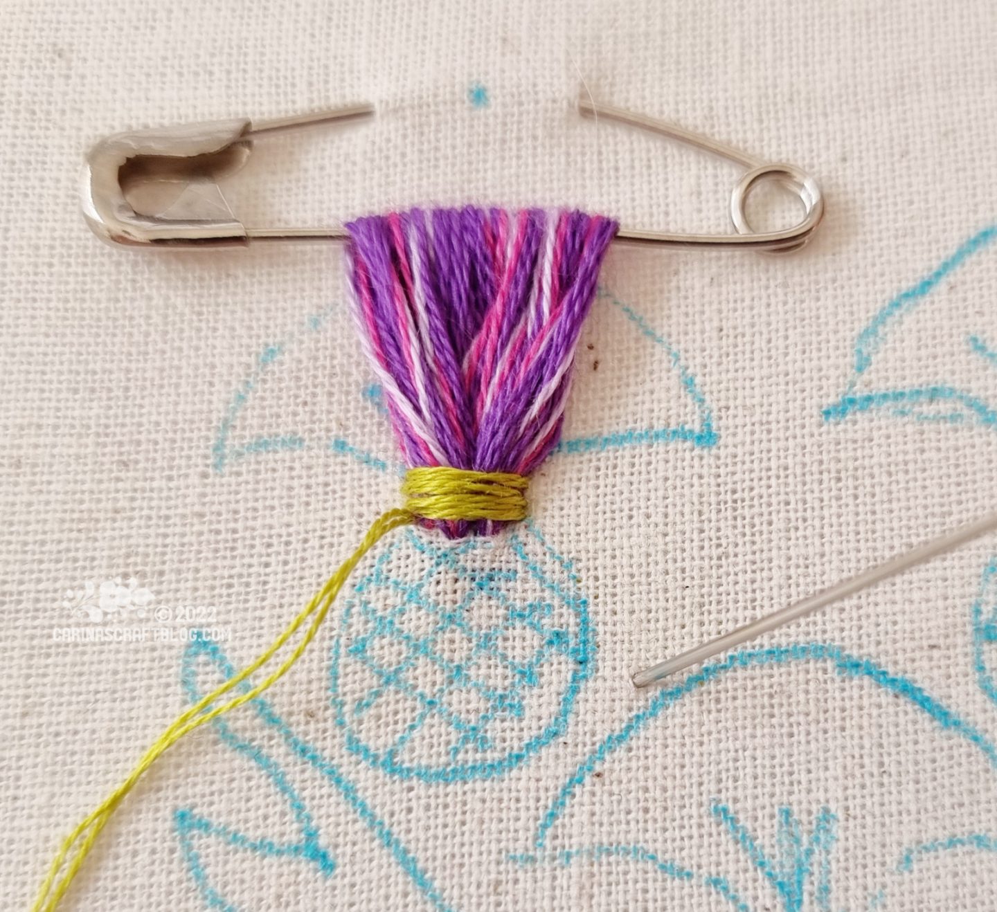 Closeup of a safety pin attached to white fabric. Embroidery thread in purple colours has been looped through the safety pin several times. At the base of the loops, green stitches are made across the pruple thread loops.