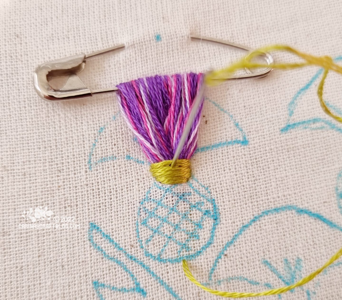 Closeup of a safety pin attached to white fabric. Embroidery thread in purple colours has been looped through the safety pin several times. At the base of the loops, green stitches are made across the purple thread loops.