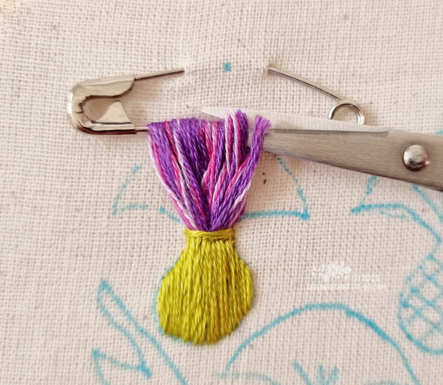 Closeup of a safety pin attached to white fabric. Embroidery thread in purple colours has been looped through the safety pin several times. At the base of the loops, green stitches are made across the purple thread loops. Scissors are shown in the process of cutting the thread loops loose from the safety pin.