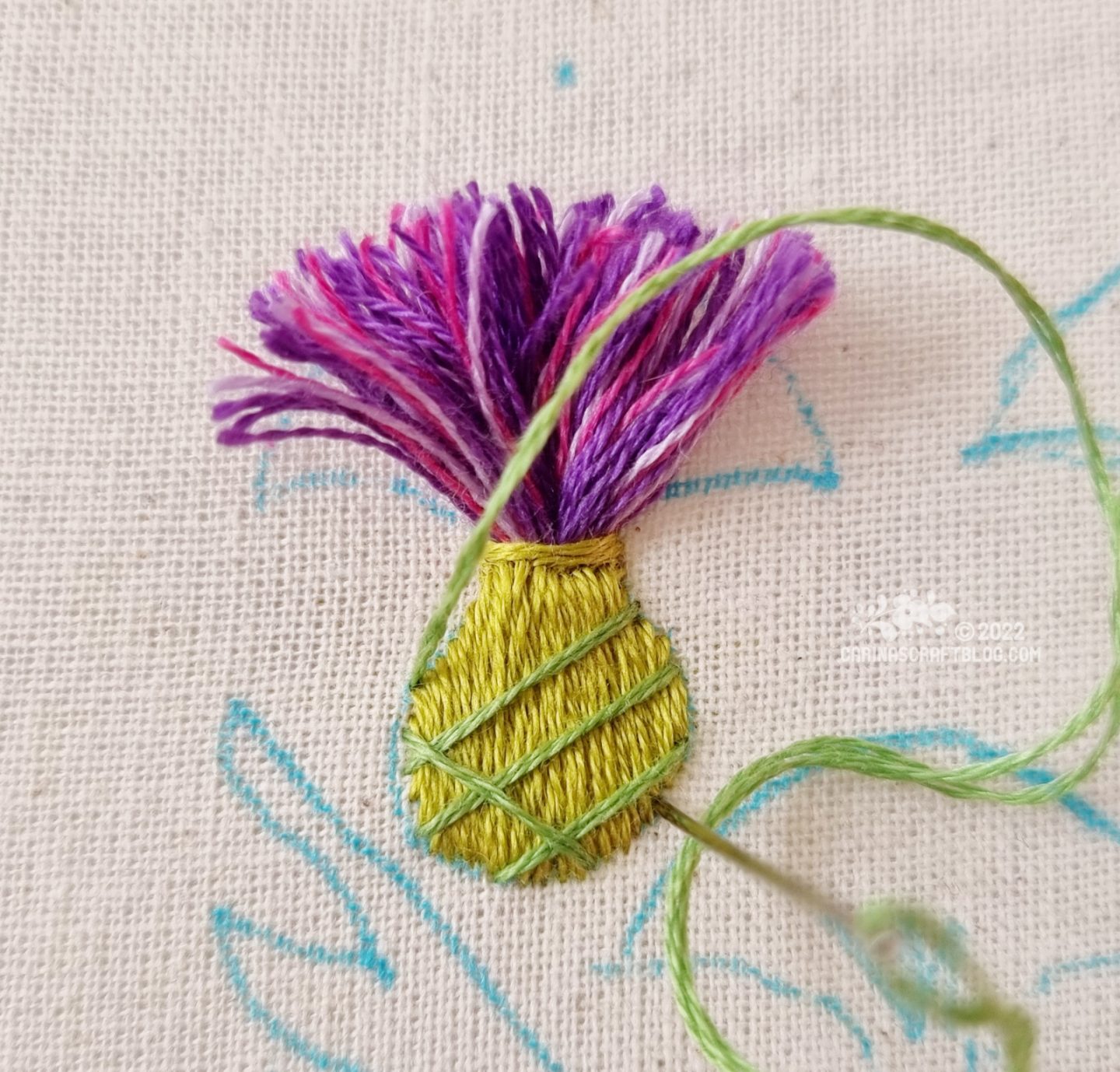 Close view of an embroidered thistle flower made from purple threads. The base is stitched with one green thread, overlaid with diagonal stitches in a different green.
