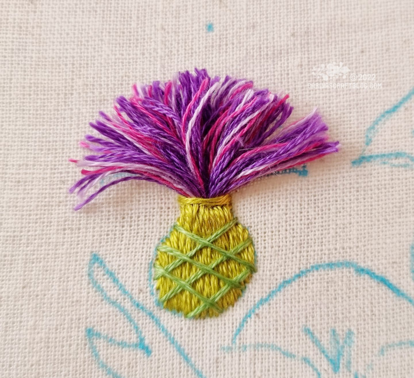 Close view of an embroidered thistle flower made from purple threads. The base is stitched with one green thread, overlaid with diagonal stitches in a different green.