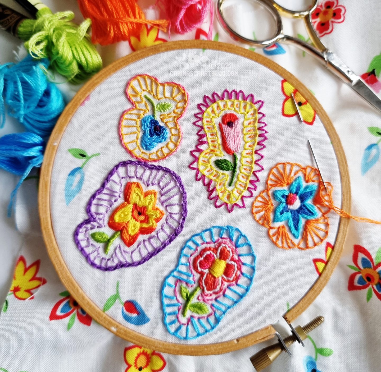 Overhead view of small embroidery hoop. In the hoop is fabric with a white base colour and flowers printed on it. Some of the flowers have been covered with hand embroidery.
