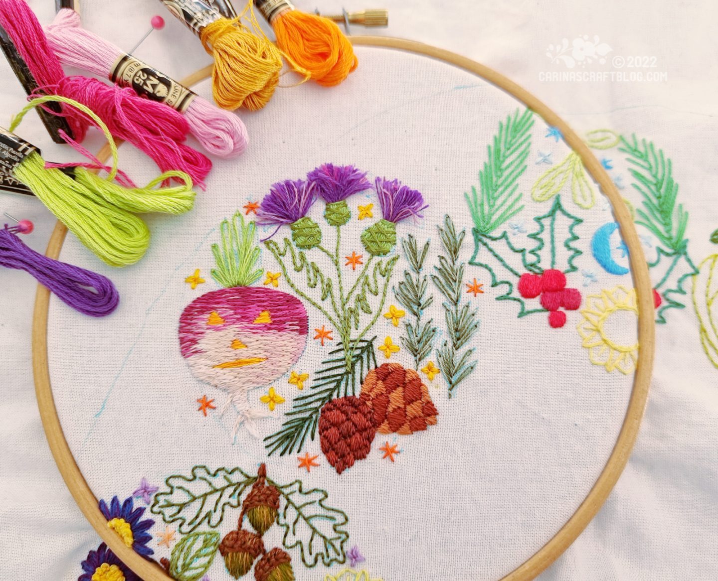 White fabric in a wood embroidery hoop. On the fabric is embroidered a design in a circular shape with thistles, pine cones, rosemary and a carved turnip.