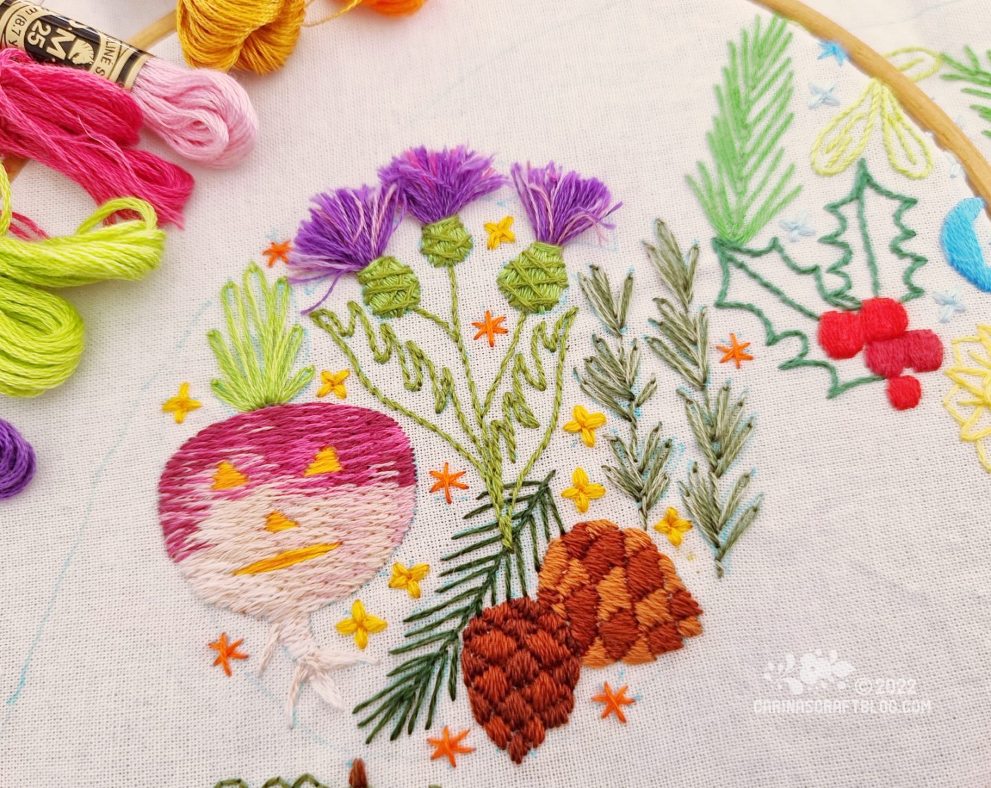 White fabric in a wood embroidery hoop. On the fabric is embroidered a design in a circular shape with thistles, pine cones, rosemary and a carved turnip.