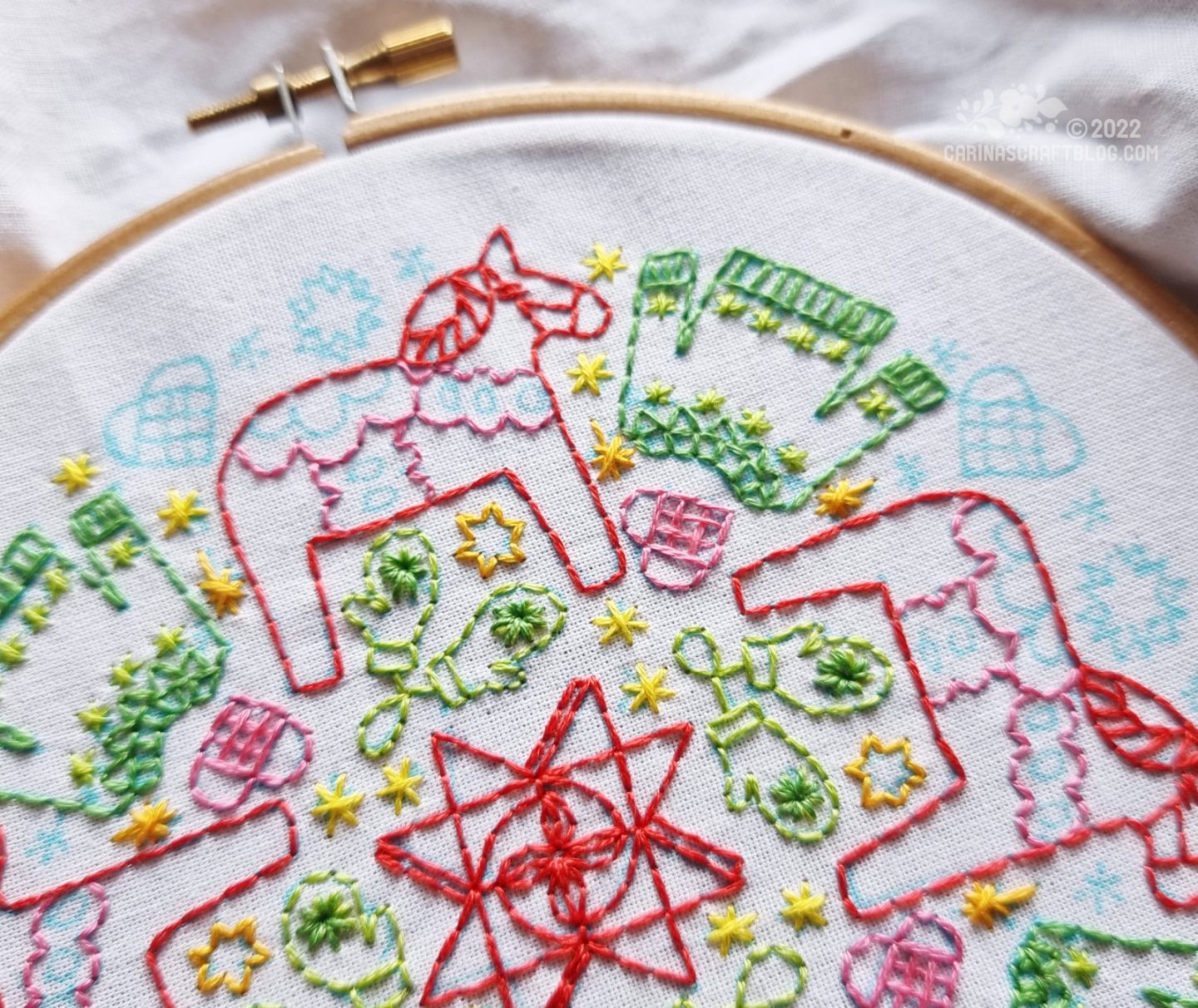 Close partial view of white fabric in a wooden embroidery hoop. On the fabric is embroidered a design with red dala horse, green sweaters, green mittens and a red woven star.