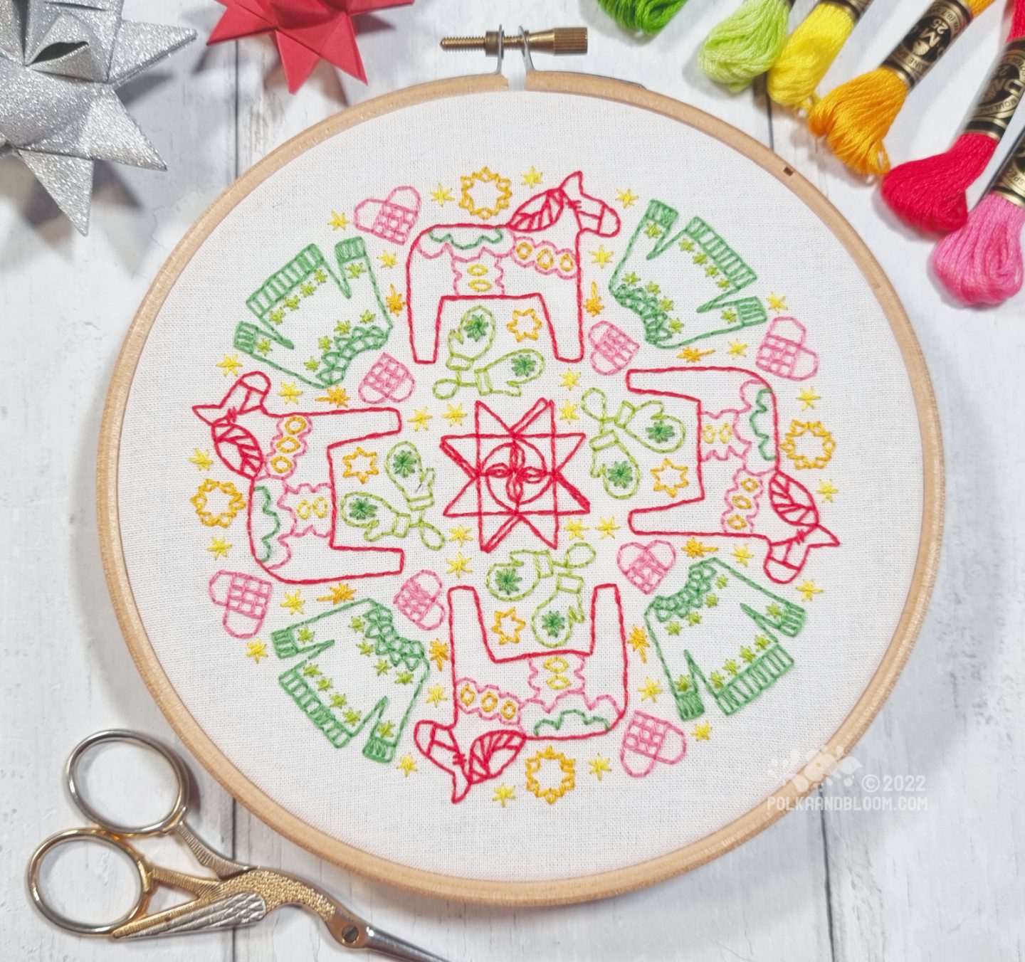 An embroidery hoop with white fabric is seen from above. On the fabric is embroidered a mandala inspired design with stars, sweaters, mittens, hears and dala horses.
