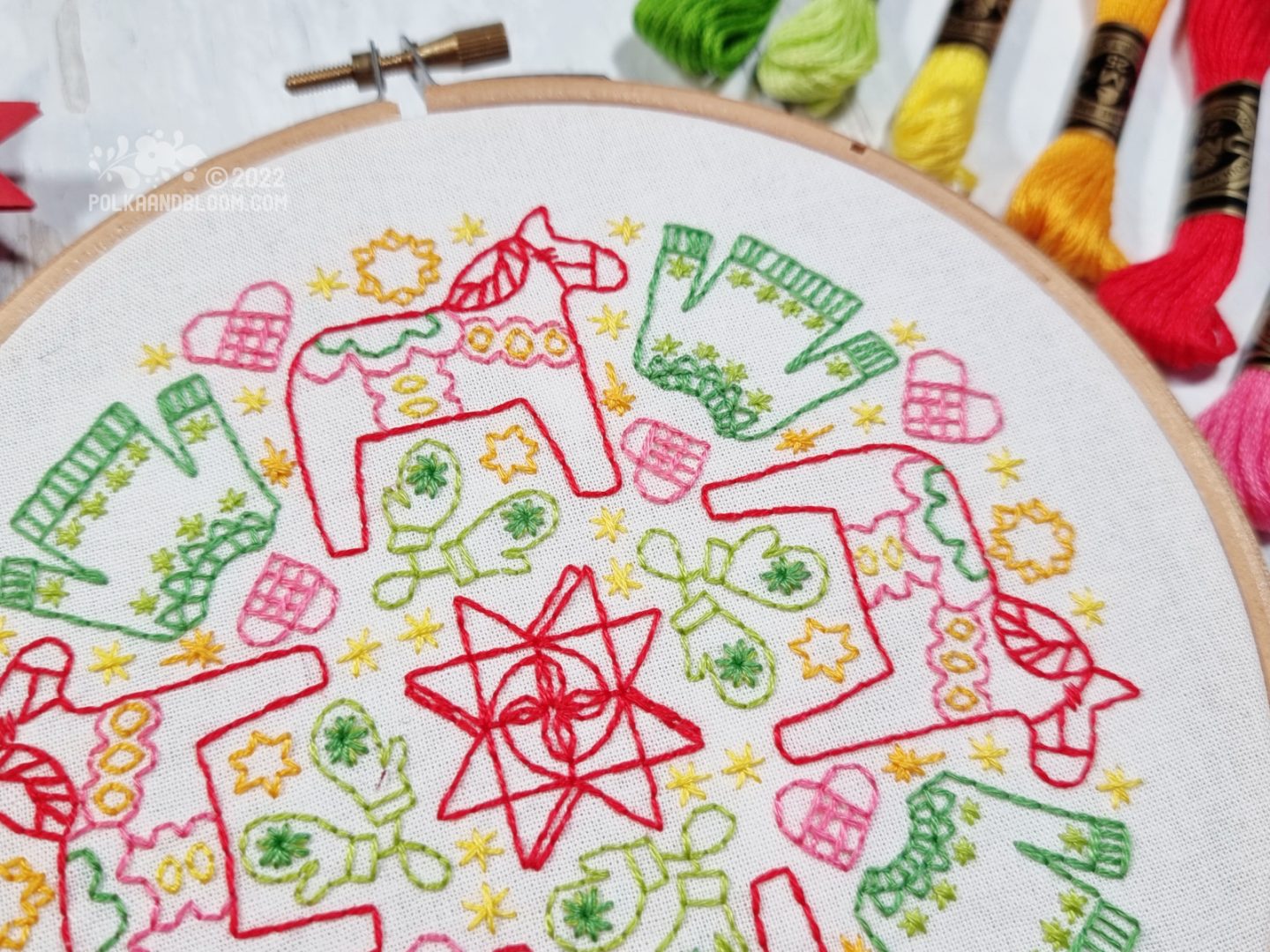 An embroidery hoop with white fabric is seen from above. On the fabric is embroidered a mandala inspired design with stars, sweaters, mittens, hears and dala horses.