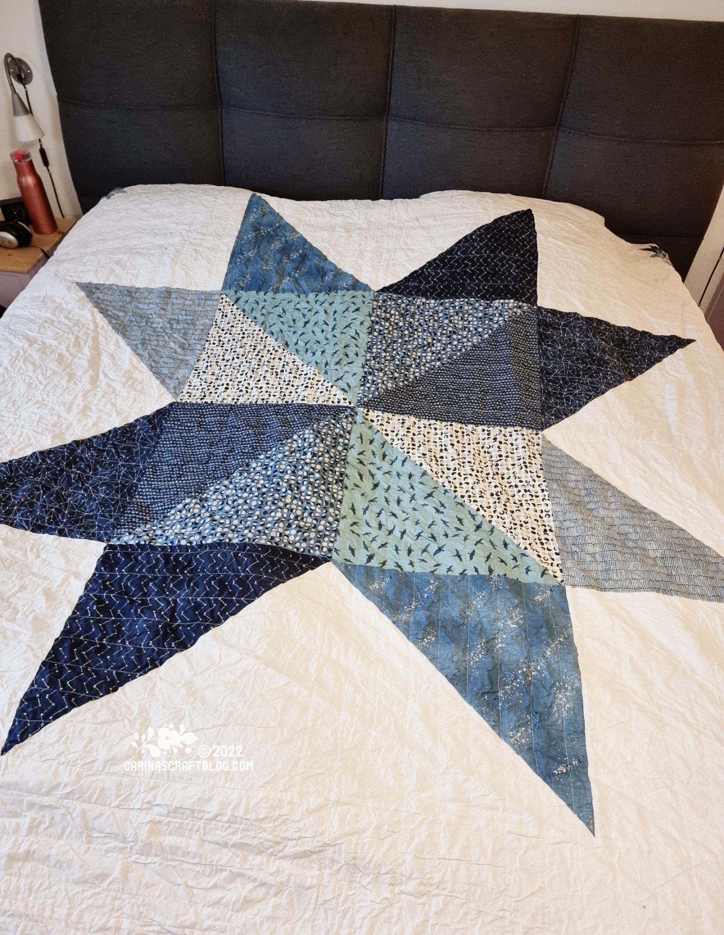 Photo of a bed with a dark grey head board. On the bed is a coverlet in eggshell white with a large star design in blue colours.