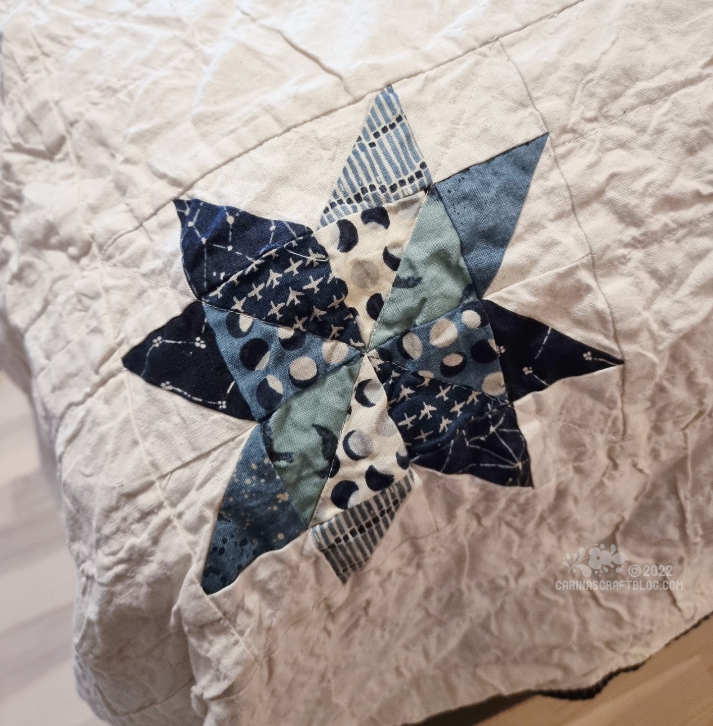 Photo of a small eight point star patchwork block in blue colours on an eggshell white background.