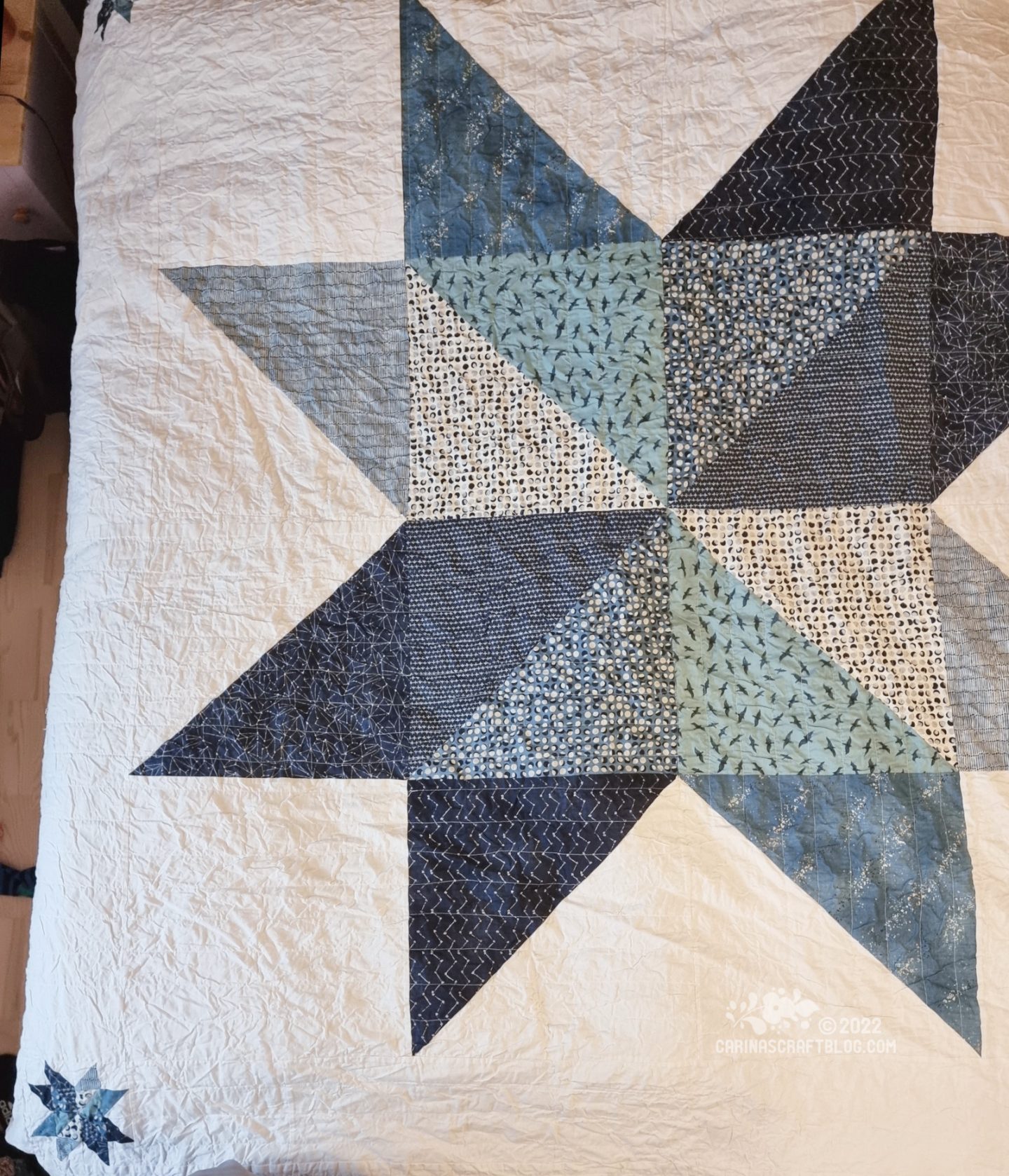 Overhead photo of a bed with a dark grey head board. On the bed is a coverlet in eggshell white with a large star design in blue colours.