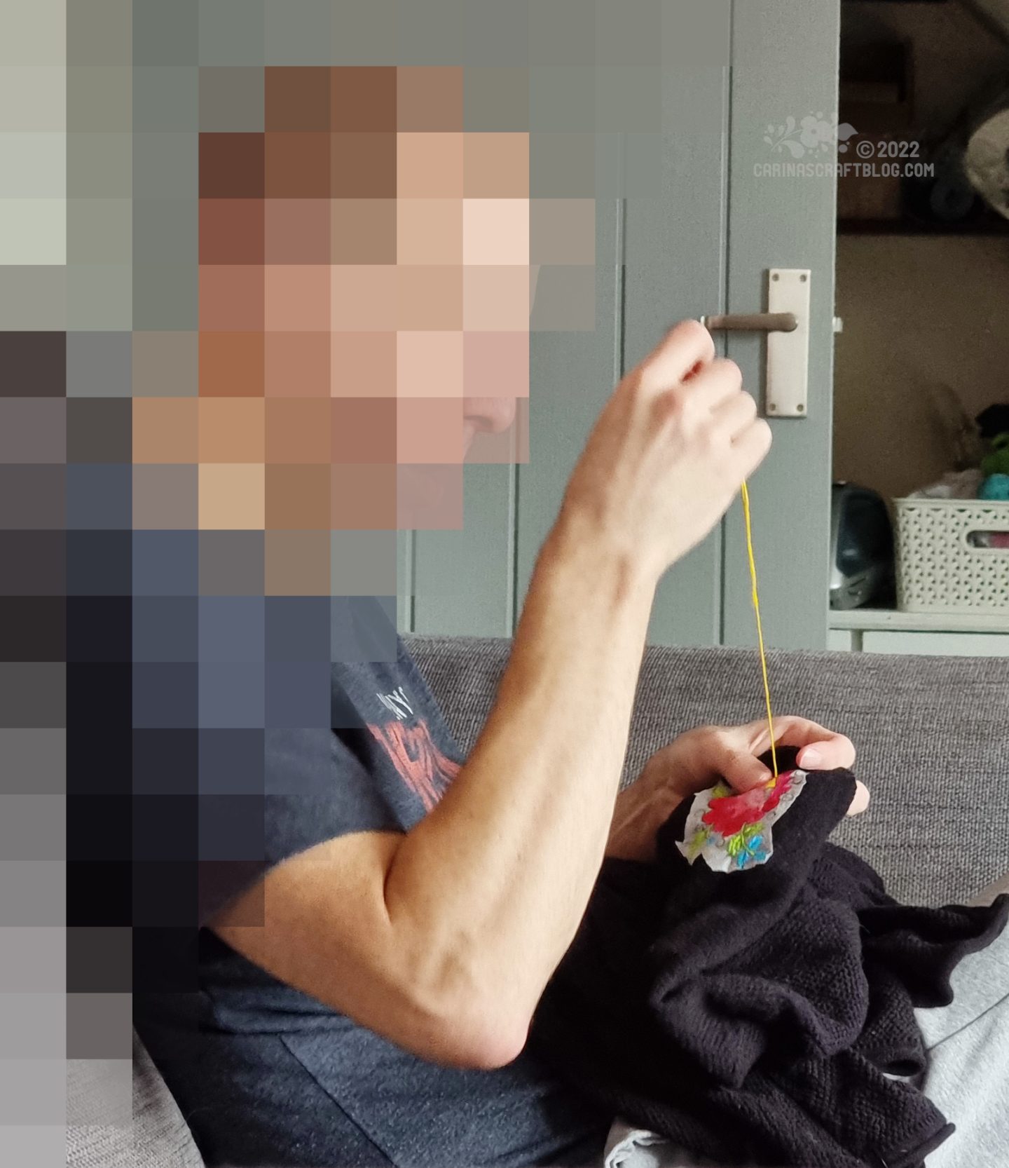 Profile view of a white man sitting on a sofa, working on embroidery on a black cardigan. The man's face is blurred with large pixels.
