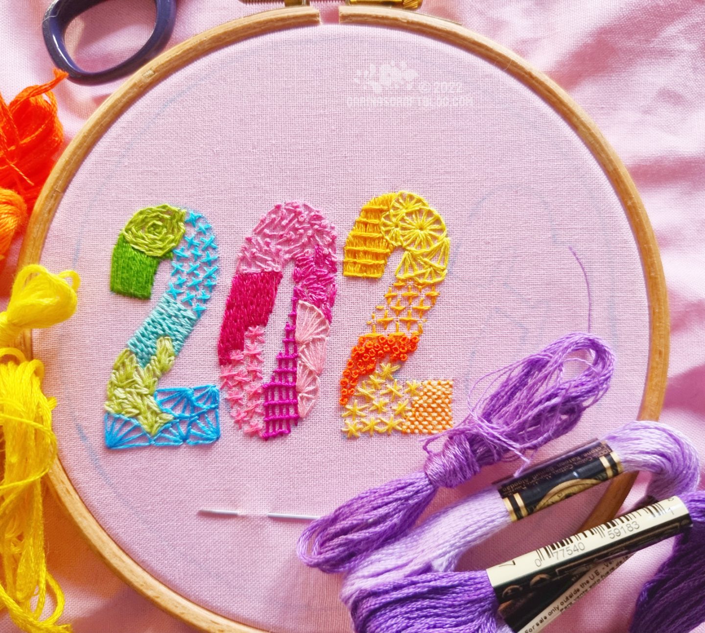 Light pink fabric in an embroidery hoop. On the fabric is drawn the number 2023. The number is filled in with various stitches in bright colours.