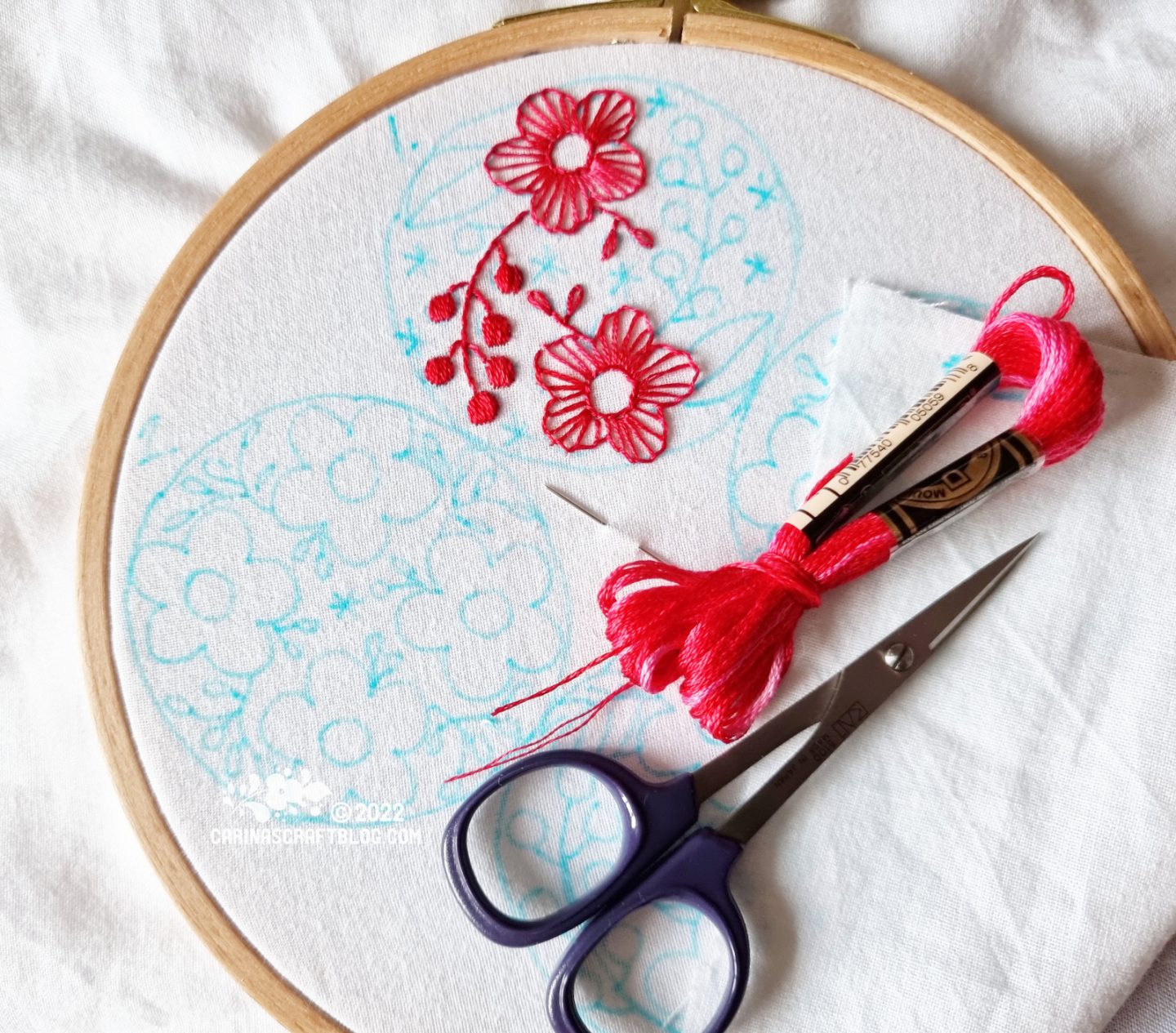 Over head view of embroidery hoop with white fabric. On the fabric is drawn two circles with flower and star motifs. One circle motif is partially stitched with dark pink thread.