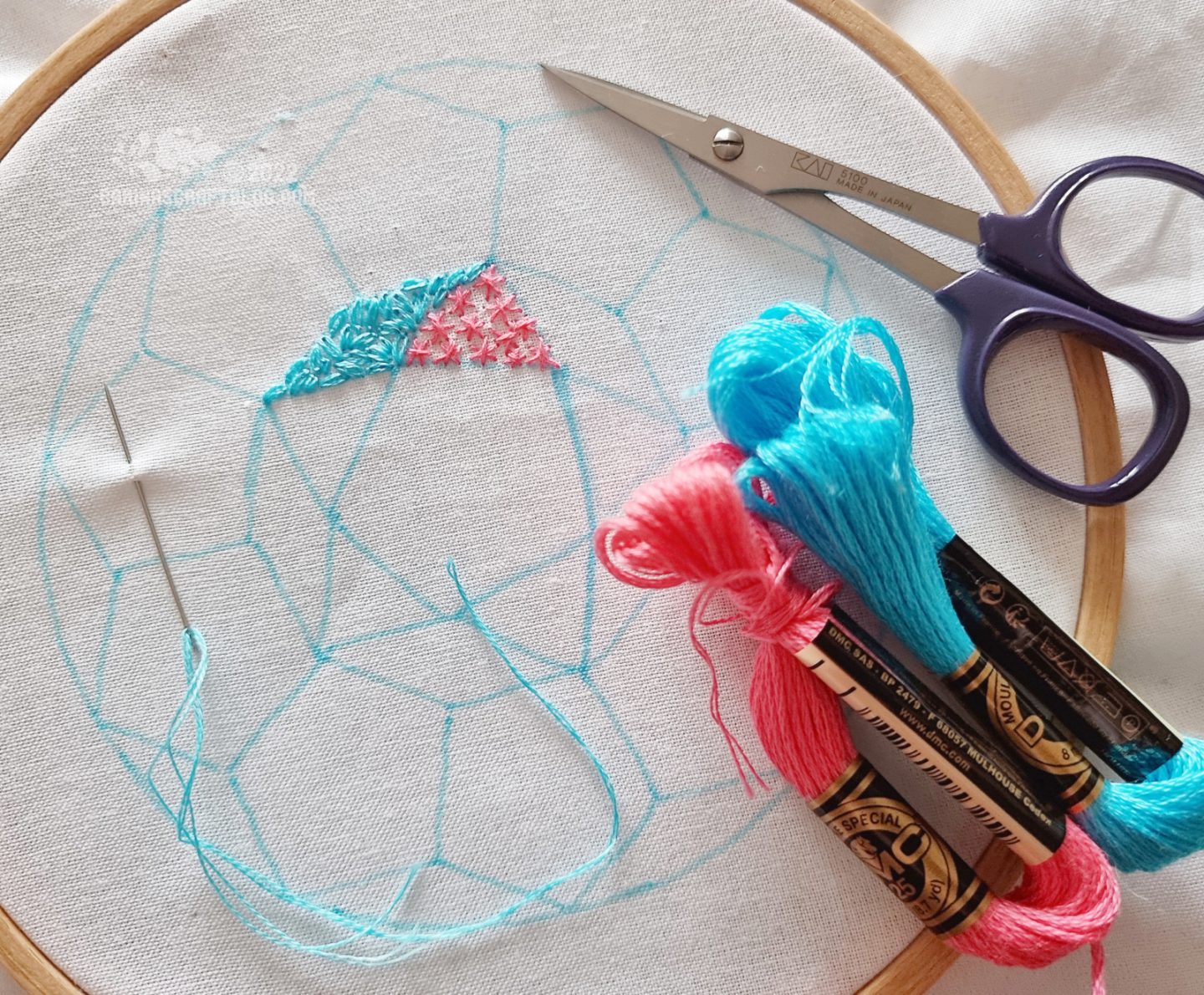 Over head view of embroidery hoop with white fabric. On the fabric is drawn a geometric design in a circle. Two of the sections are filled with stitches.