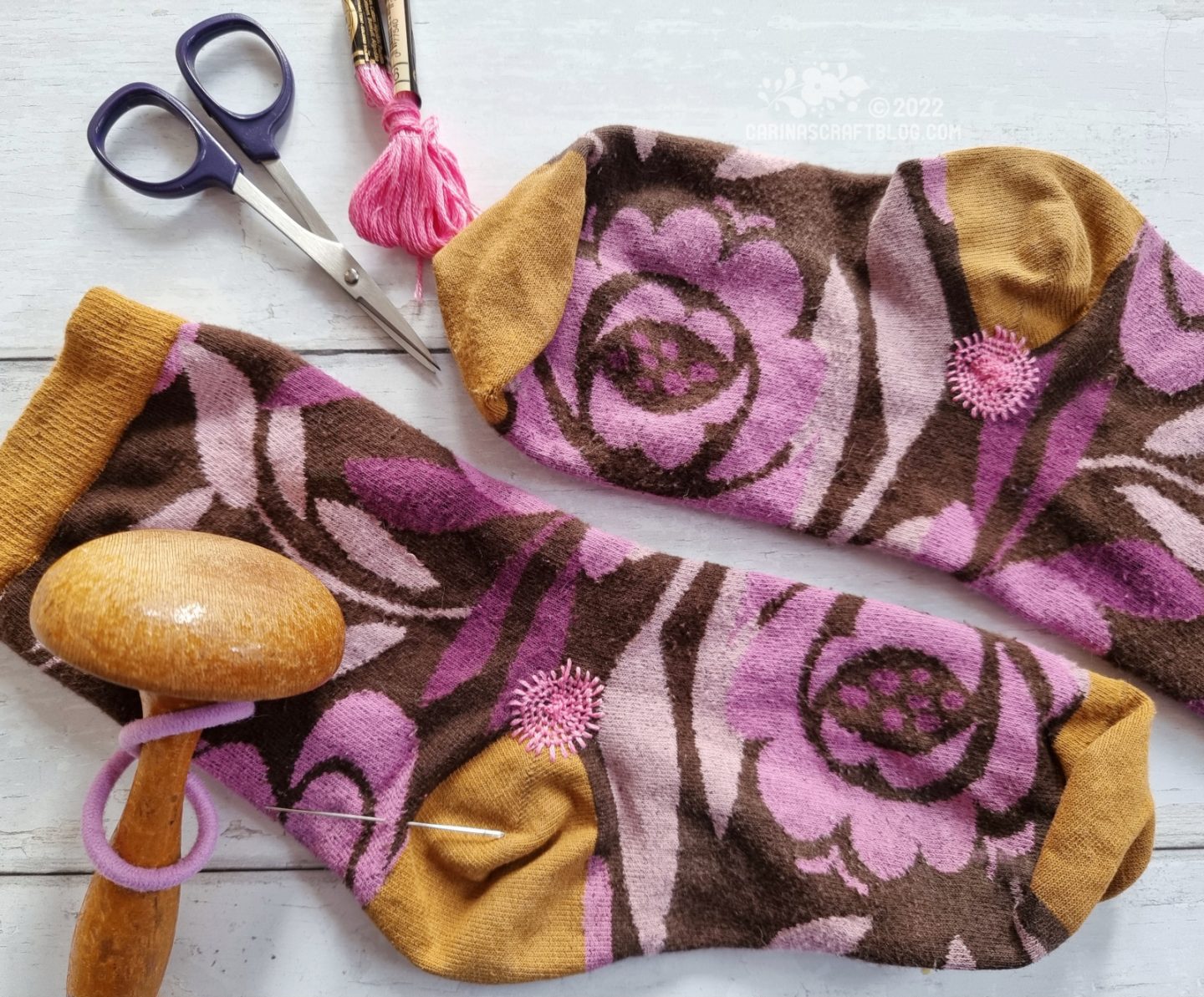 A pair of brown socks with a pink flower pattern and mustard coloured heels and toes. Surrounded by skein of pink thread, a small pair of scissors and a wooden darning mushroom.