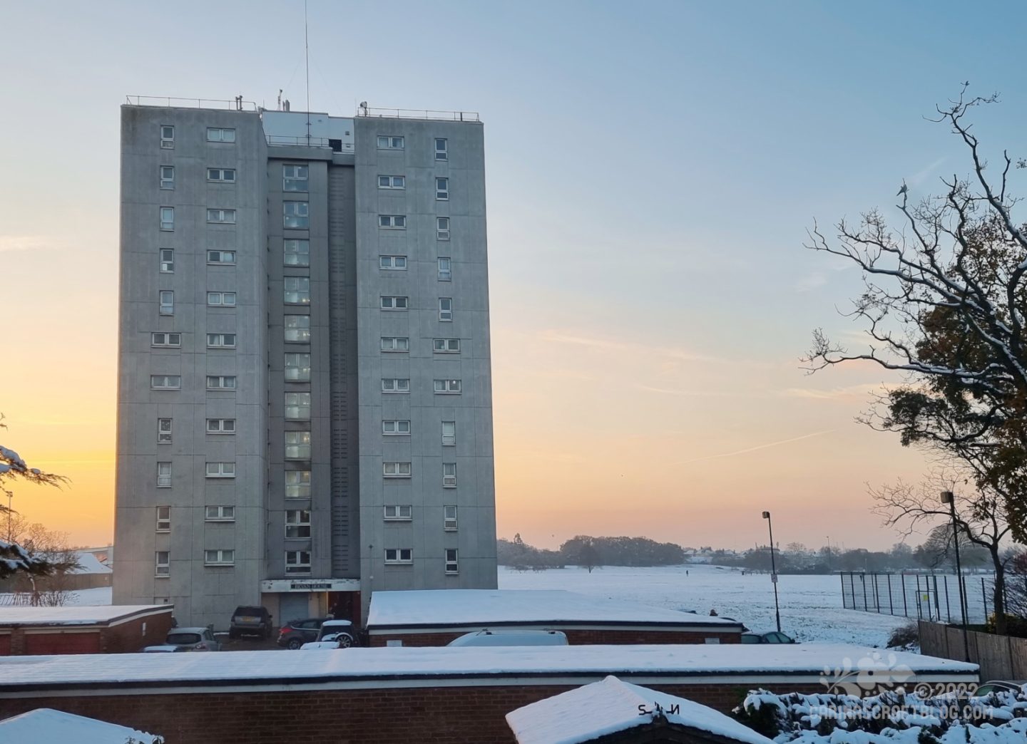 A block of flats stands in front of a snow covered field. In front of  the flats are rows of garages with snow covered roofs. In the back ground the sky is an orange ombre colour from the rising sun.