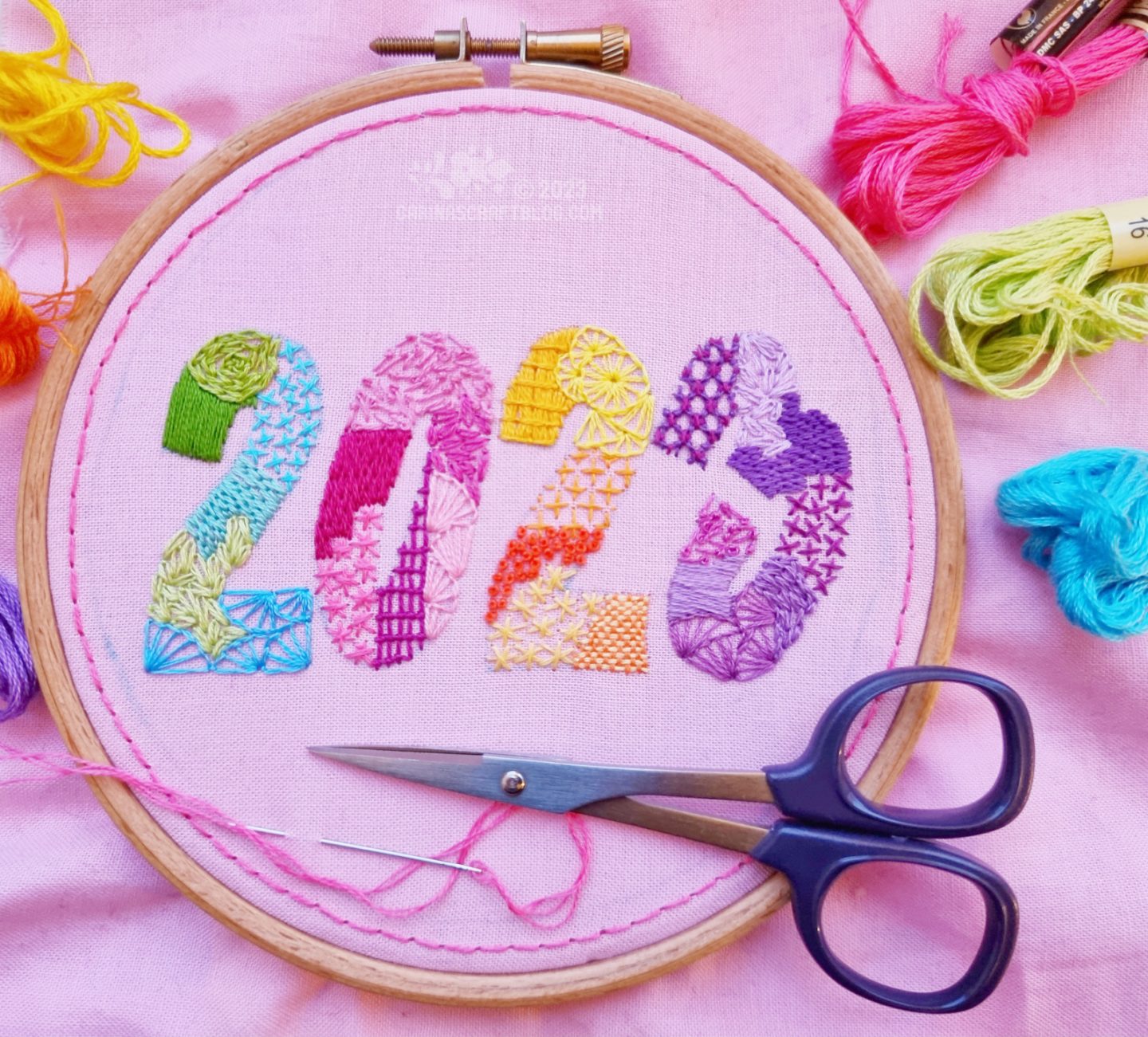 Light pink fabric in a wooden embroidery hoop. On the fabric is embroidered the number 2023 using many different stitches and colours.