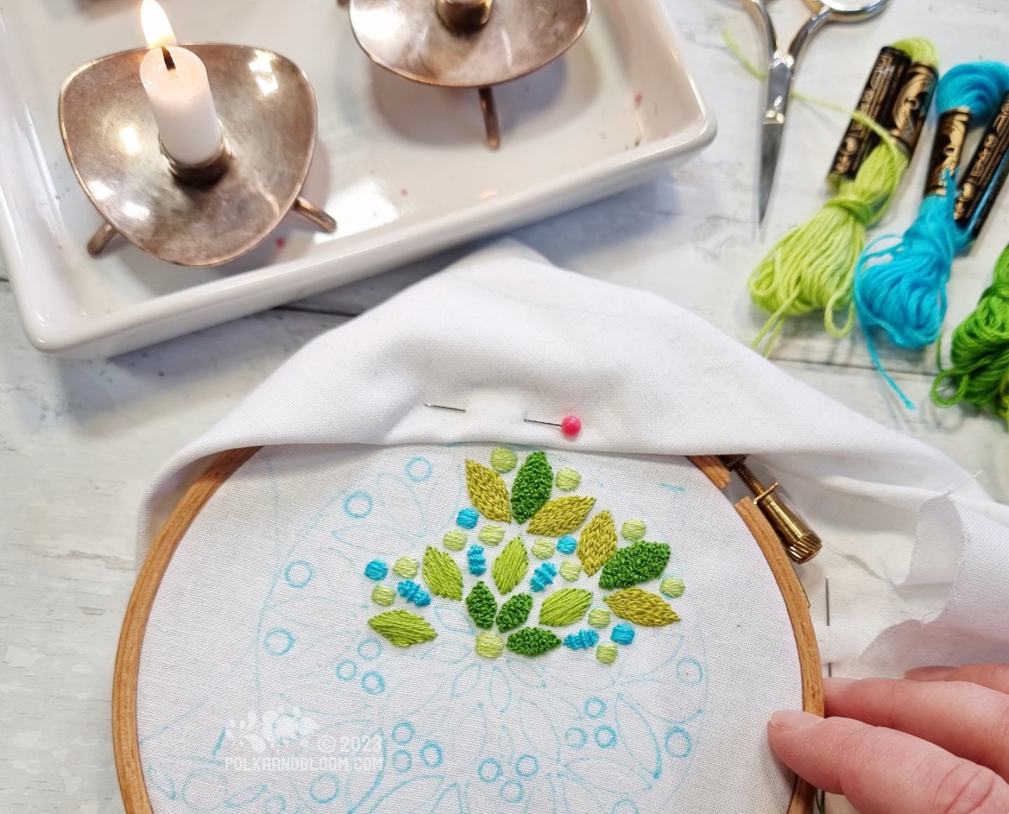 Overhead shot of an embroidery hoop with white fabric. A mandala style design is partially embroidered on the fabric with green and aqua colour threads.