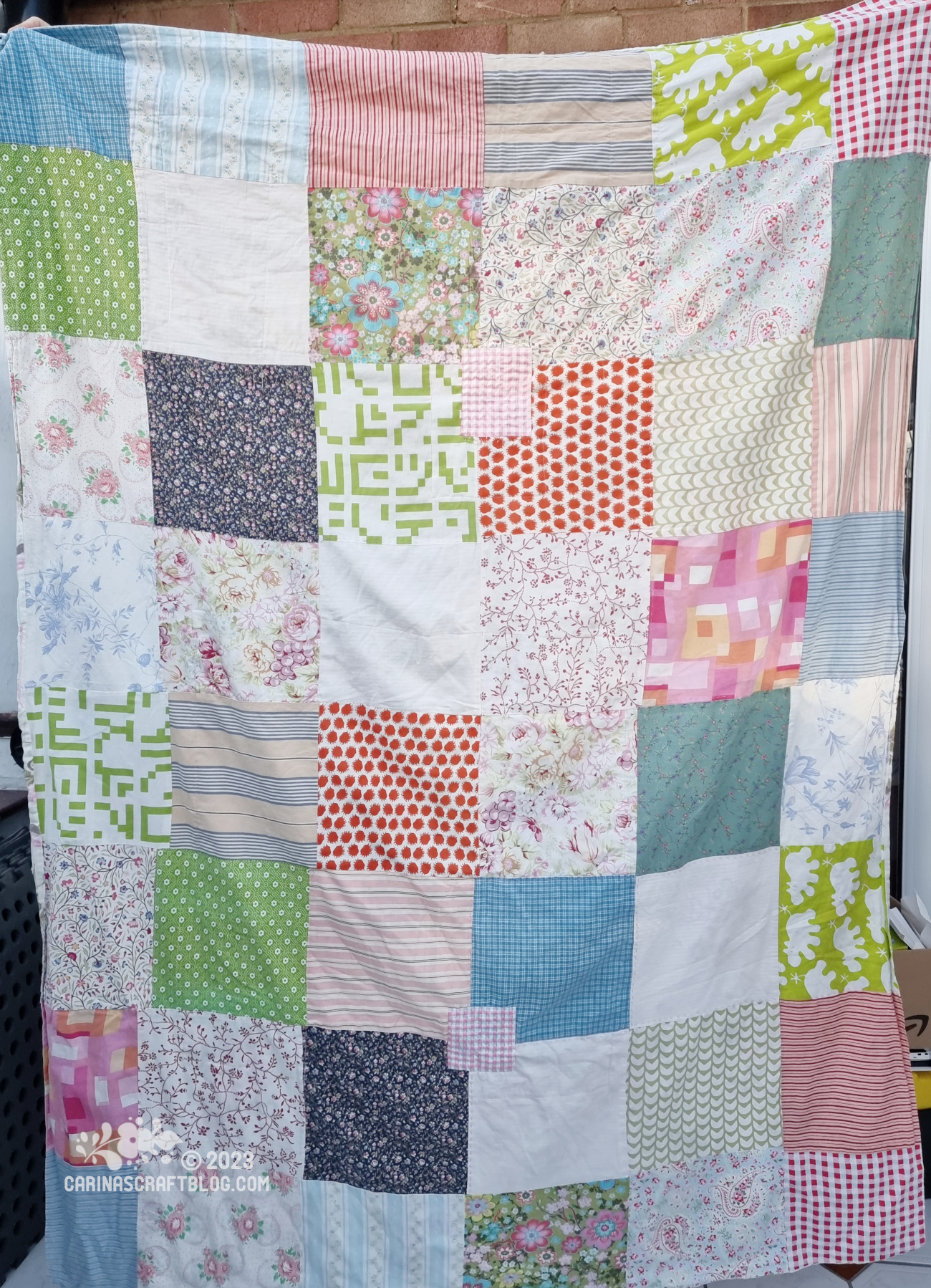 The front of a fabric blanket made up of square patches in different colours and patterns, mostly red, green, blue and taupe colours.