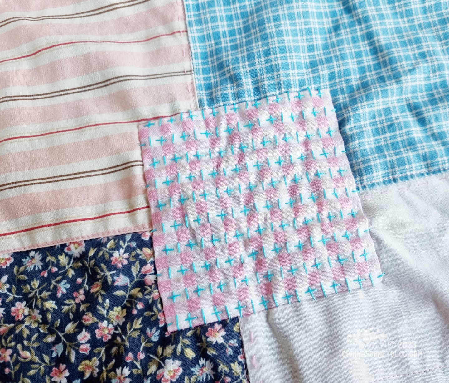 Close view of a mended patch in a pink and white check pattern on a patchwork blanket. Blue hand stitches on top of the patch.