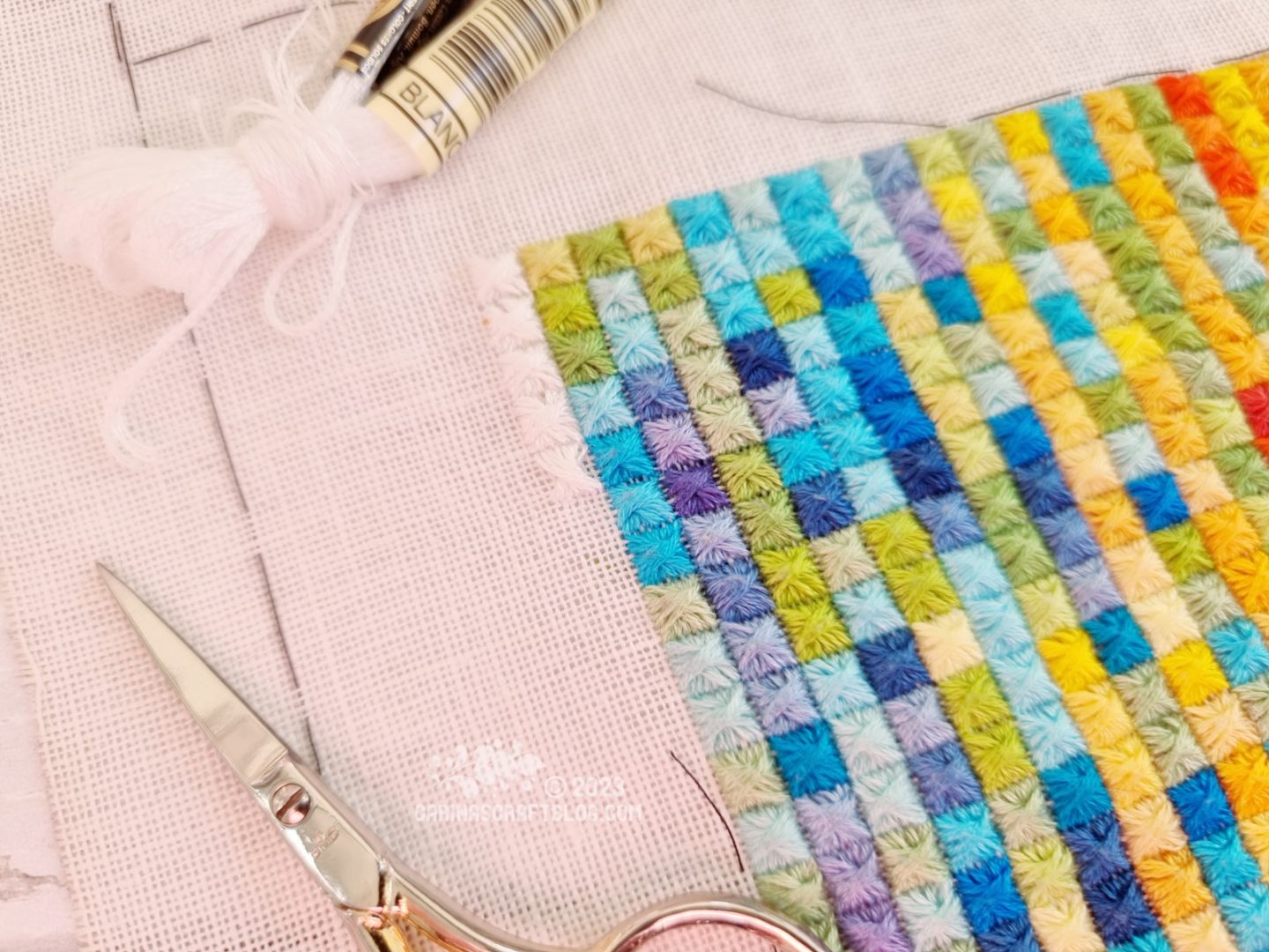 Closeup view of a multicolour embroidery made up of rows and columns of square stitches. Along the top left corner are white square stitches.