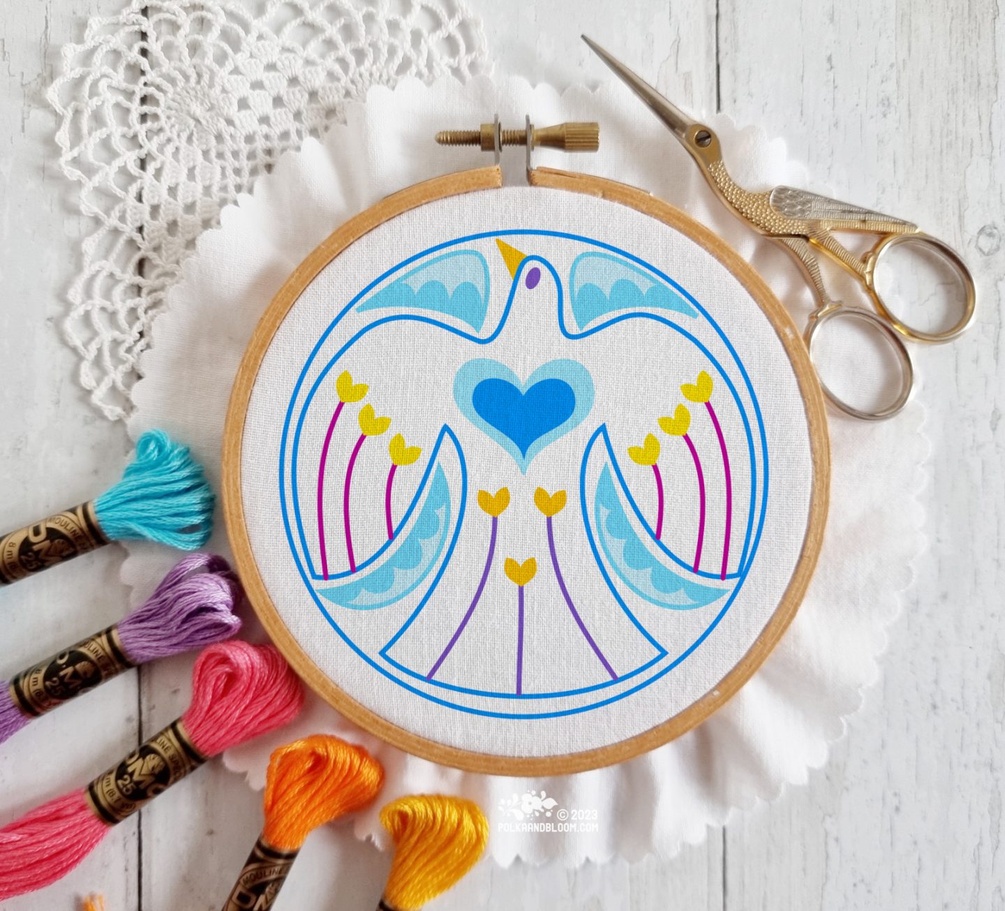 An illustration of a blue bird in a blue circle is inserted on a photo of an embroidery hoop with white fabric, surrounded by skeins of embroidery thread.