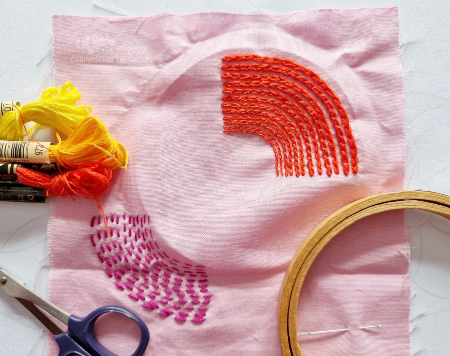 A light pink square of fabric is stitched in two rainbow shapes. An orange shape in the upper right corner and a magenta shape in the lower left corner.