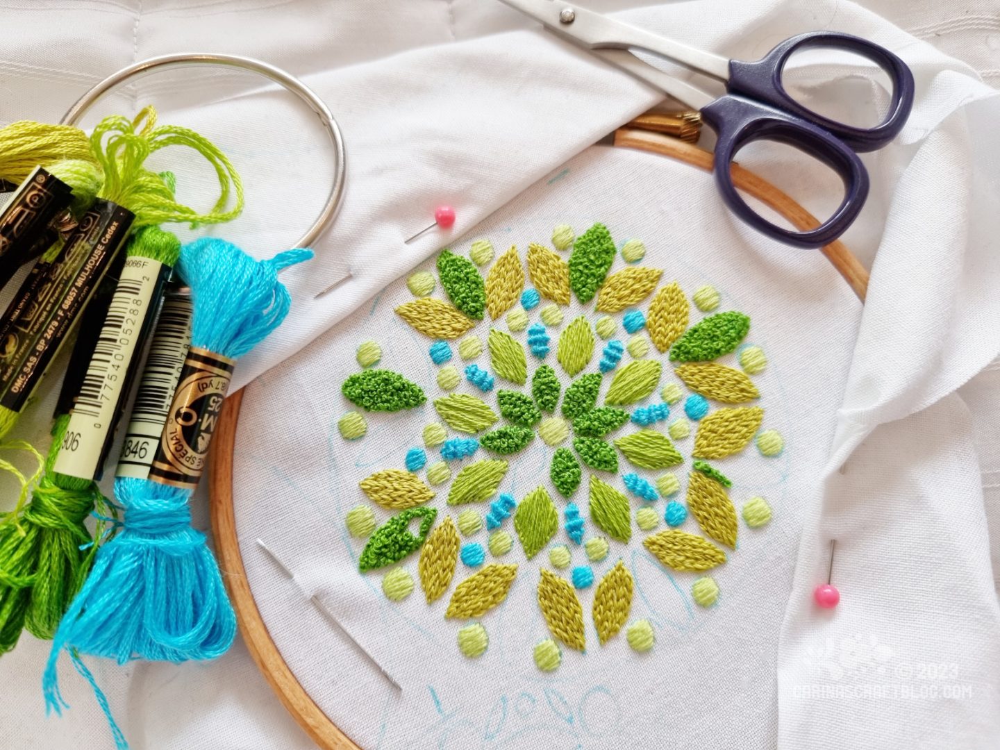 A mandala inspired design is partially stitched in green and turquoise colours on white fabric stretched in a wooden embroidery hoop.