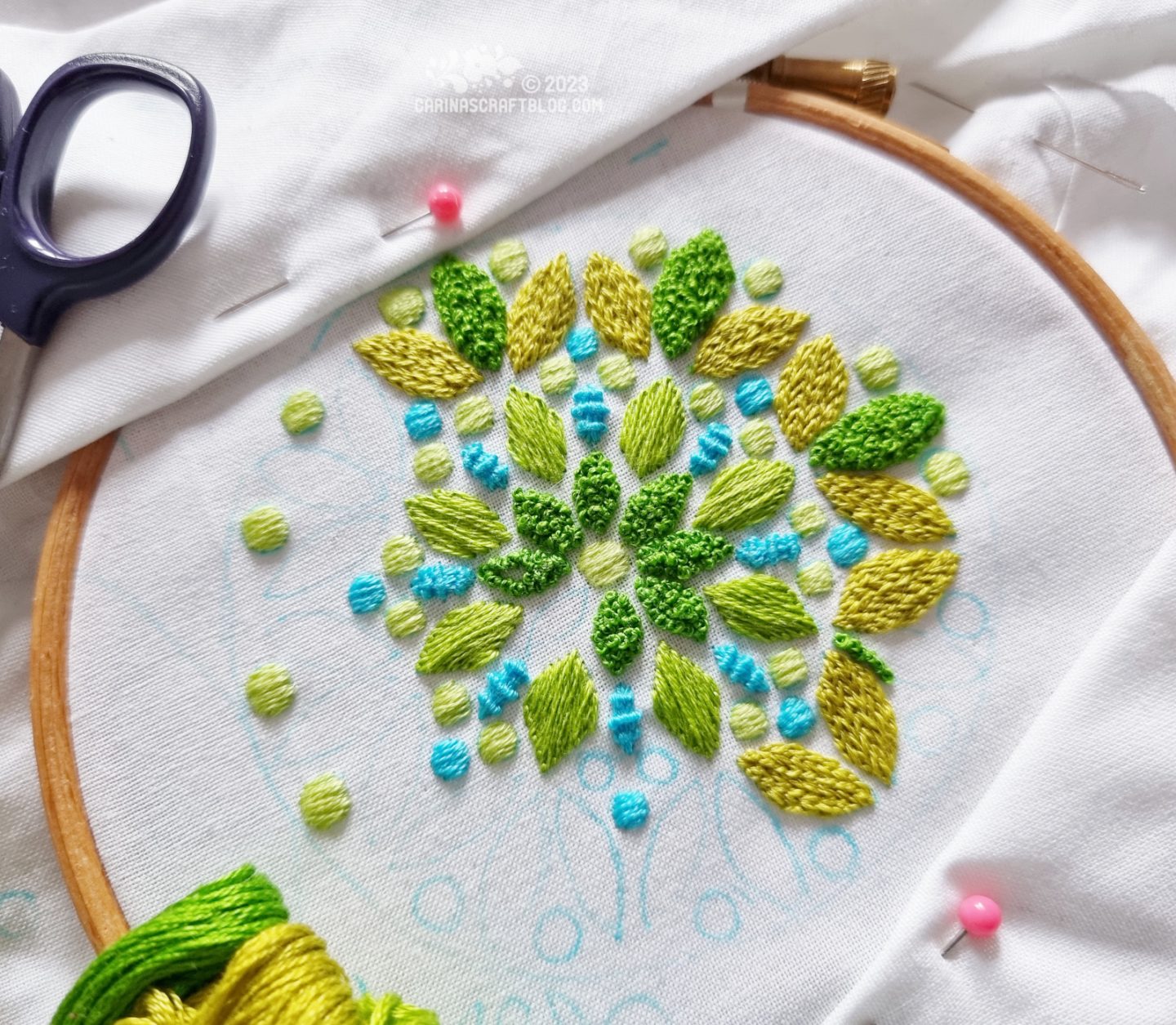 White fabric in a small wooden embroidery hoop. On the fabric is a partially embroidered mandala inspired design in green.