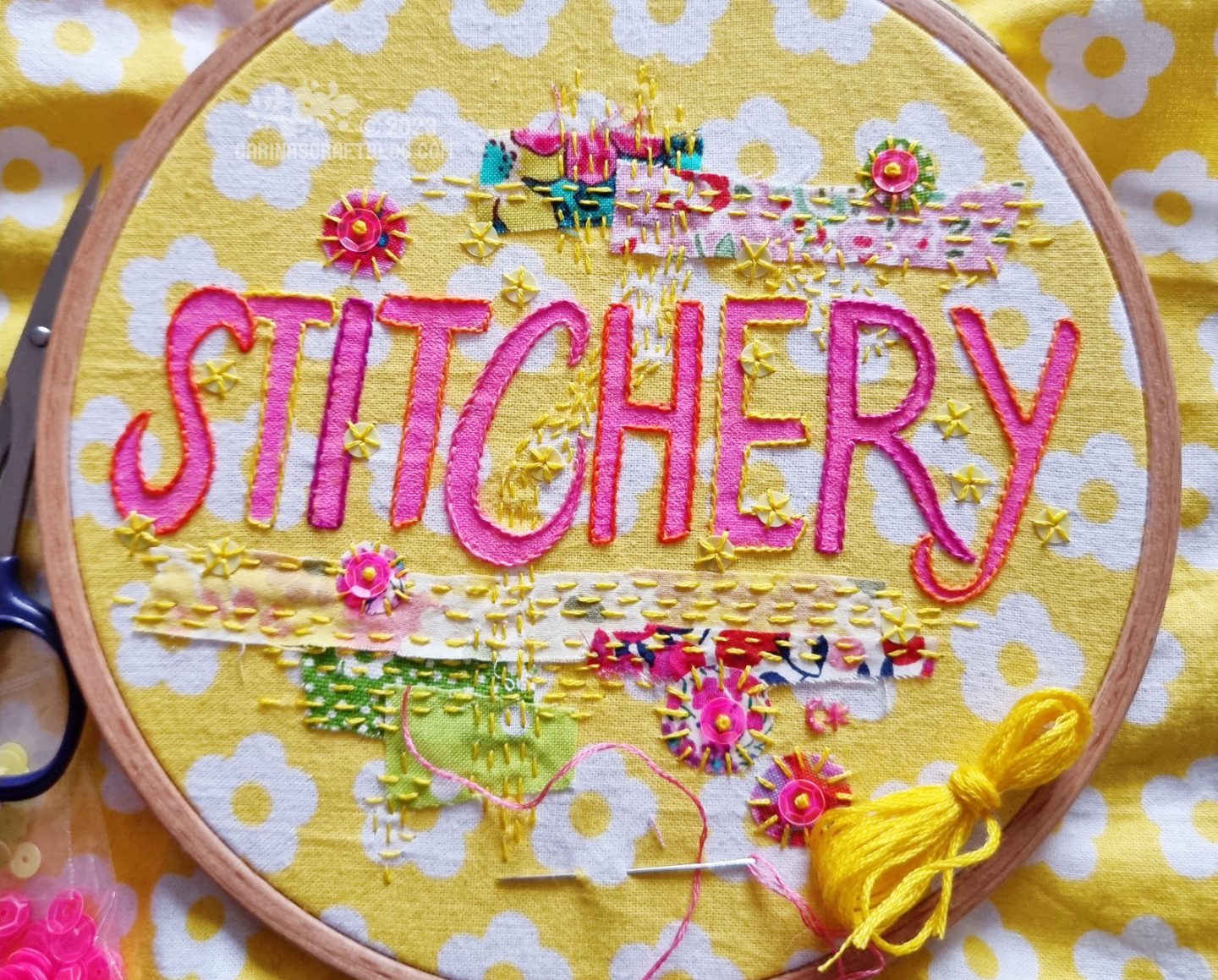 Yellow fabric with white flowers stretched in a wooden embroidery hoop. On the fabric is drawn the word stitchery in pink. Small patches of fabric in pink and green are attached to the fabric with stitches in yellow. There are pink and yellow sequins as embellishments.