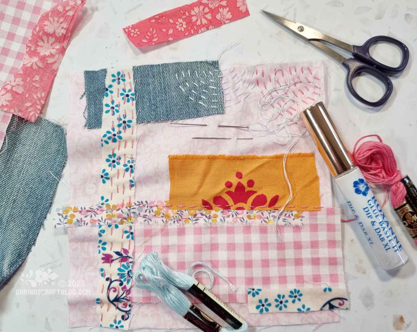 A pale pink square of fabric has been partially covered in various fabric scraps and running stitches. The square is surrounded by various threads and a pair of embroidery scissors.