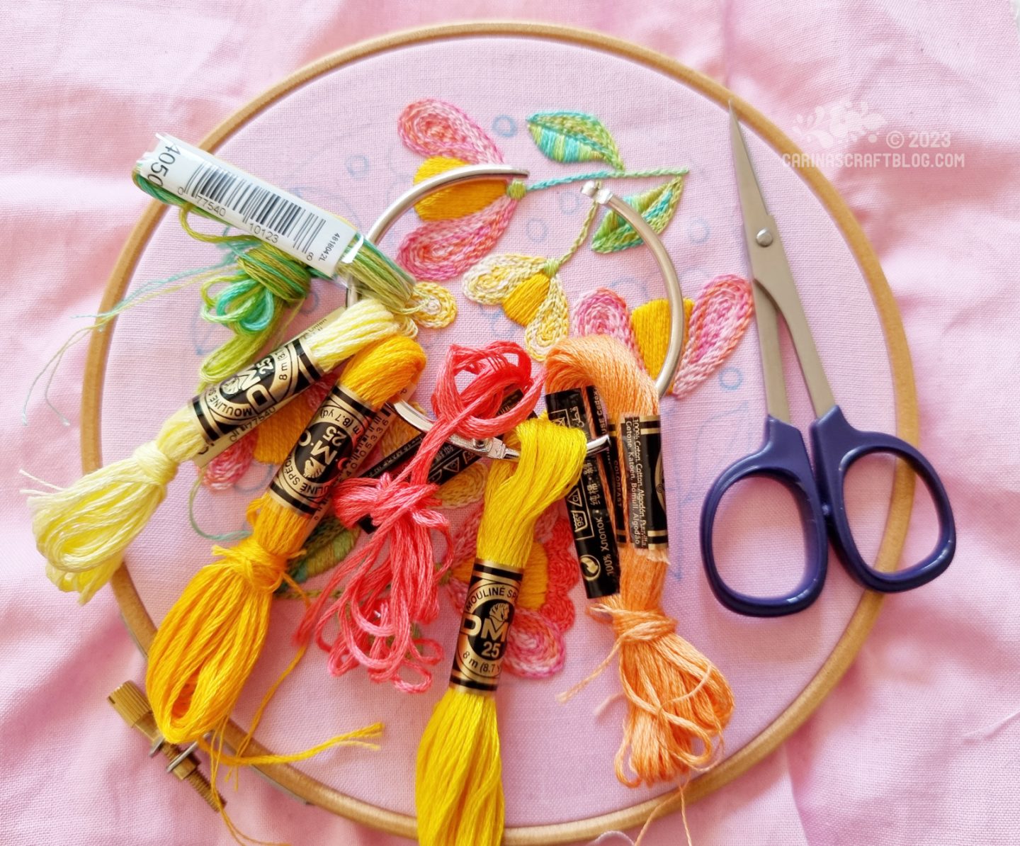 In the background is an embroidery hoop with pink fabric. On top of the fabric is a pair of embroidery scissors and a book binding ring with several skeins of embroidery thread attached to it.