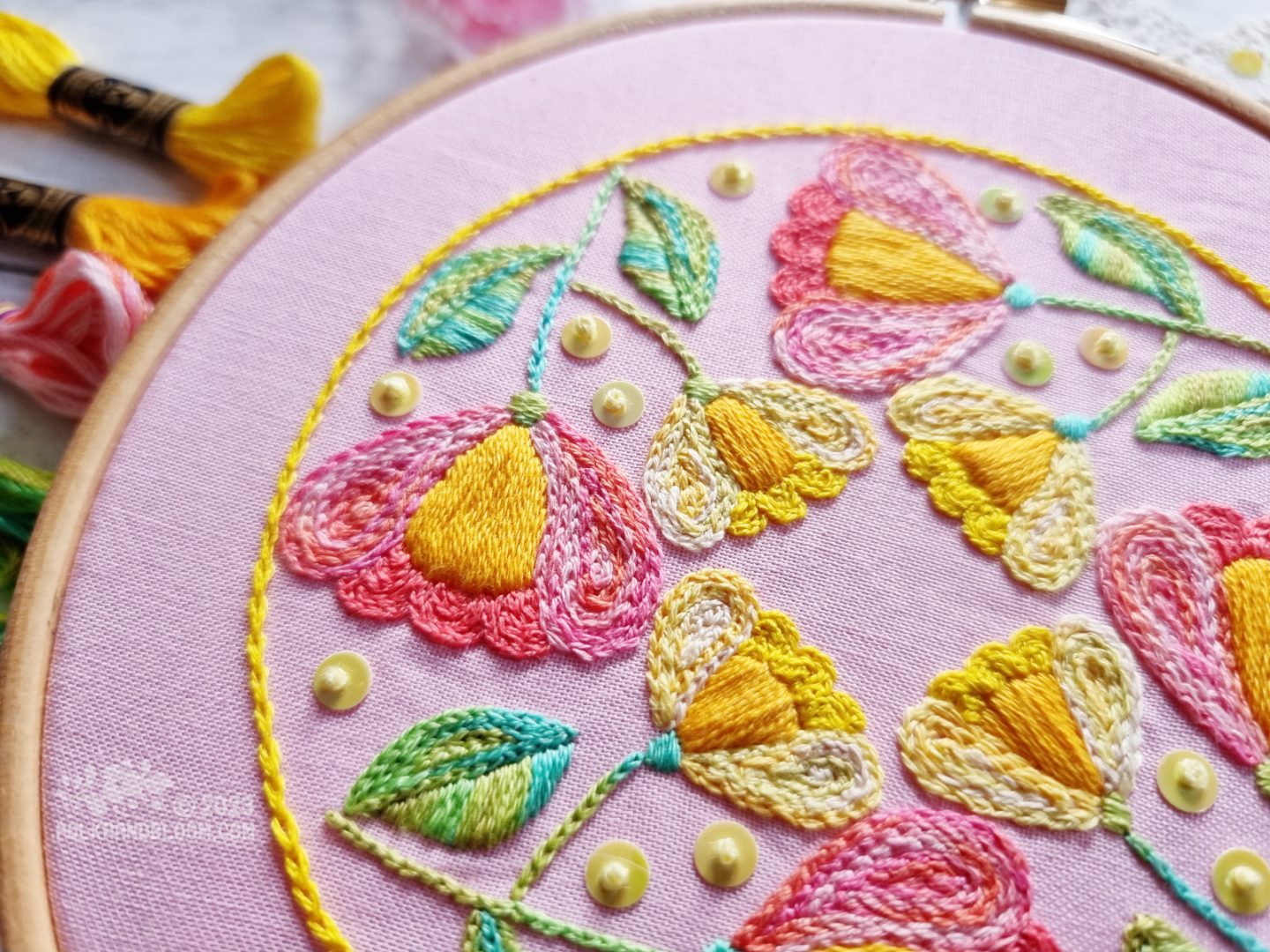 Close up view of an embroidery hoop with light pink fabric. On the fabric is stitched a circular motif with a yellow outline and four folk art inspired tulips inside the outline.