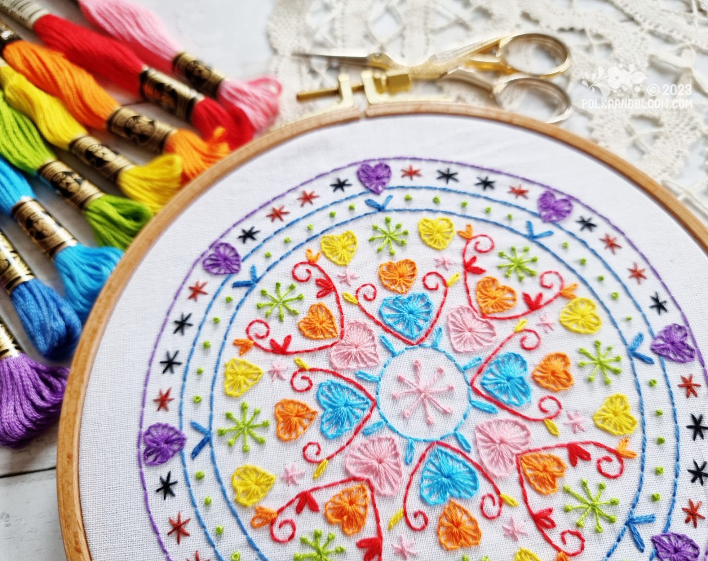 Partial view of an embroidery hoop with white fabric. On the fabric is embroidered a mandala inspired design with colours inspired by the Progress Pride flag.
