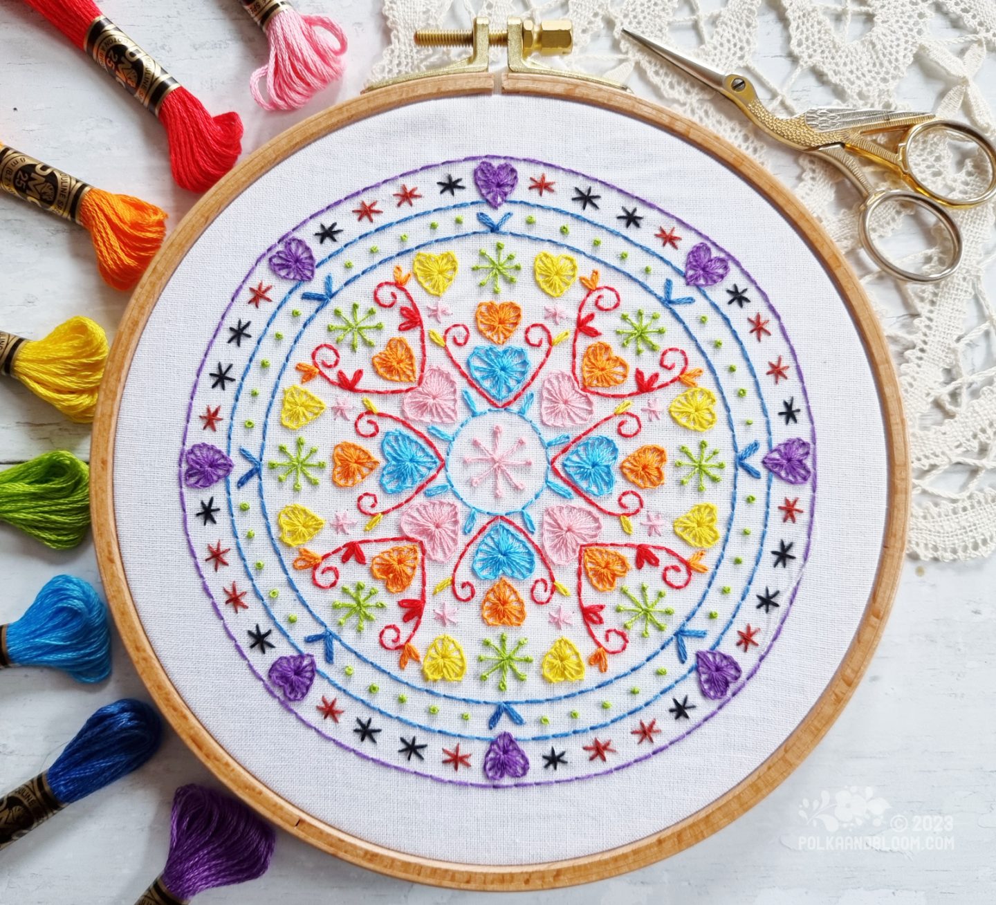 Overhead view of an embroidery hoop with white fabric. On the fabric is embroidered a mandala inspired design with colours inspired by the Progress Pride flag.