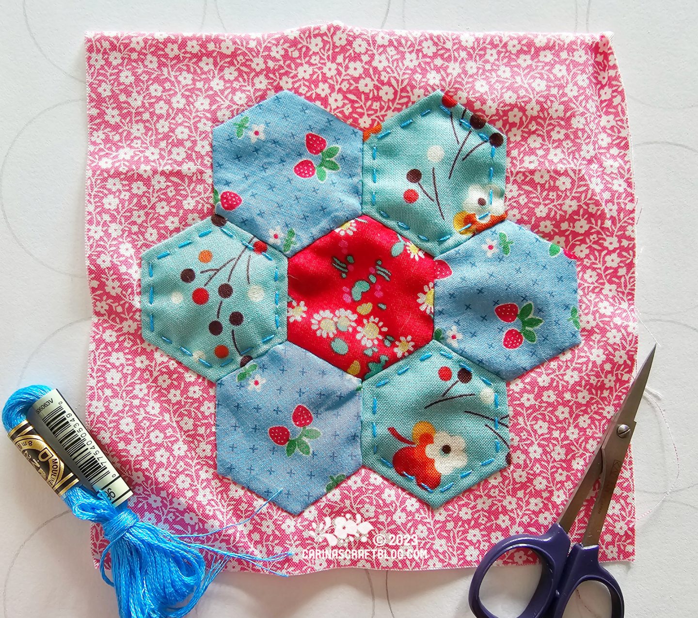 A pink square of fabric with a print of tiny white flowers. On the fabric are appliquéd seven fabric hexagons, a red hexagon in the centre with alternating blue and turquoise hexagons around it.