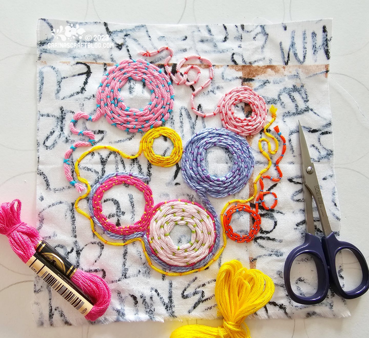 A white square of fabric with black pattern seen from above. On the fabric are embroidered circles in yellow, pink and lilac colours in varying thicknesses of thread.