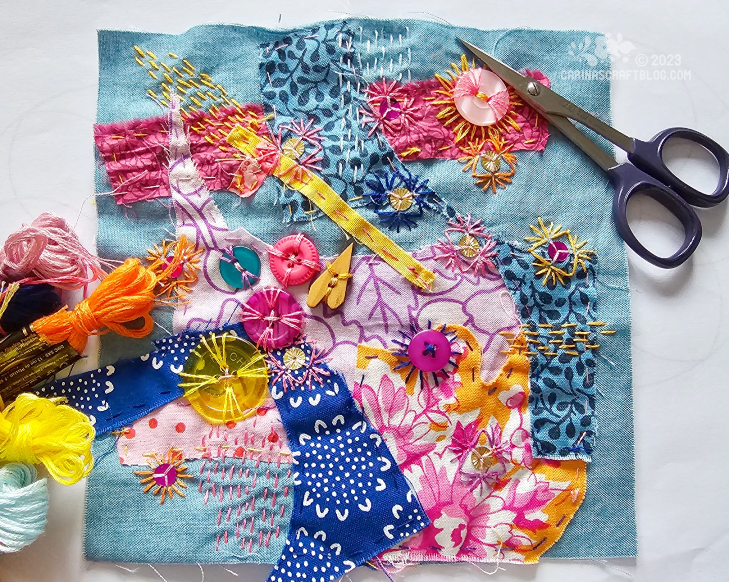 A blue square of fabric partially covered in various fabric scraps, buttons, sequins and stitches in mostly pink, orange, yellow and blue colours.