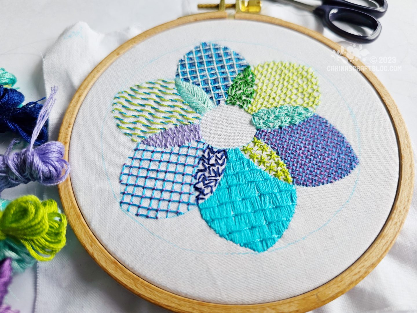 A wooden embroidery hoop with white fabric. On the fabric is embroidered a flower shaped design in cool colours using 12 different stitches.