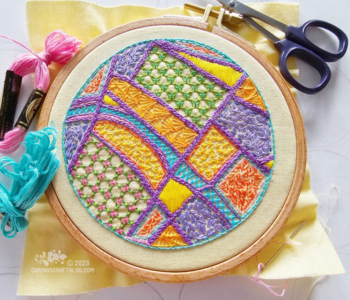 A wooden embroidery hoop with yellow fabric. On the fabric is embroidered a a map with purple thread for the roads and yellow, orange,, lilac and green colours fill in the areas between the roads.