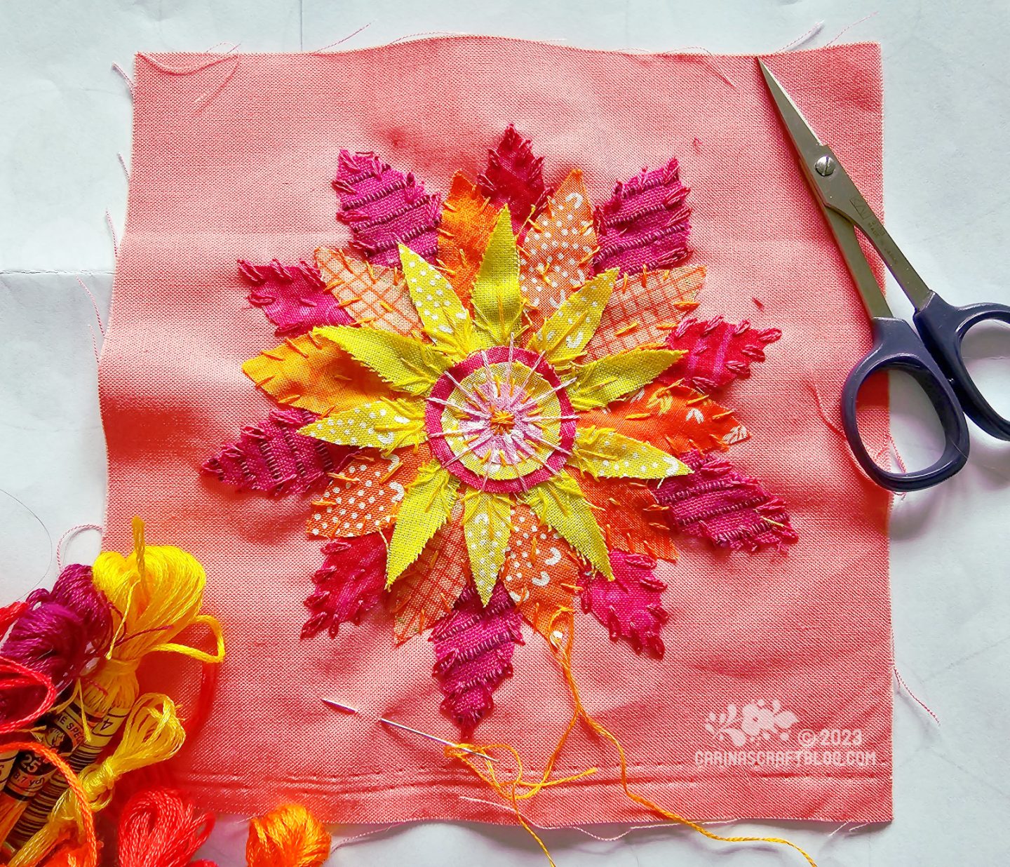 A salmon coloured square of fabric seen from above. On the fabric are appliquéd pieces of fabric in magenta, orange and yellow to form a flower. 